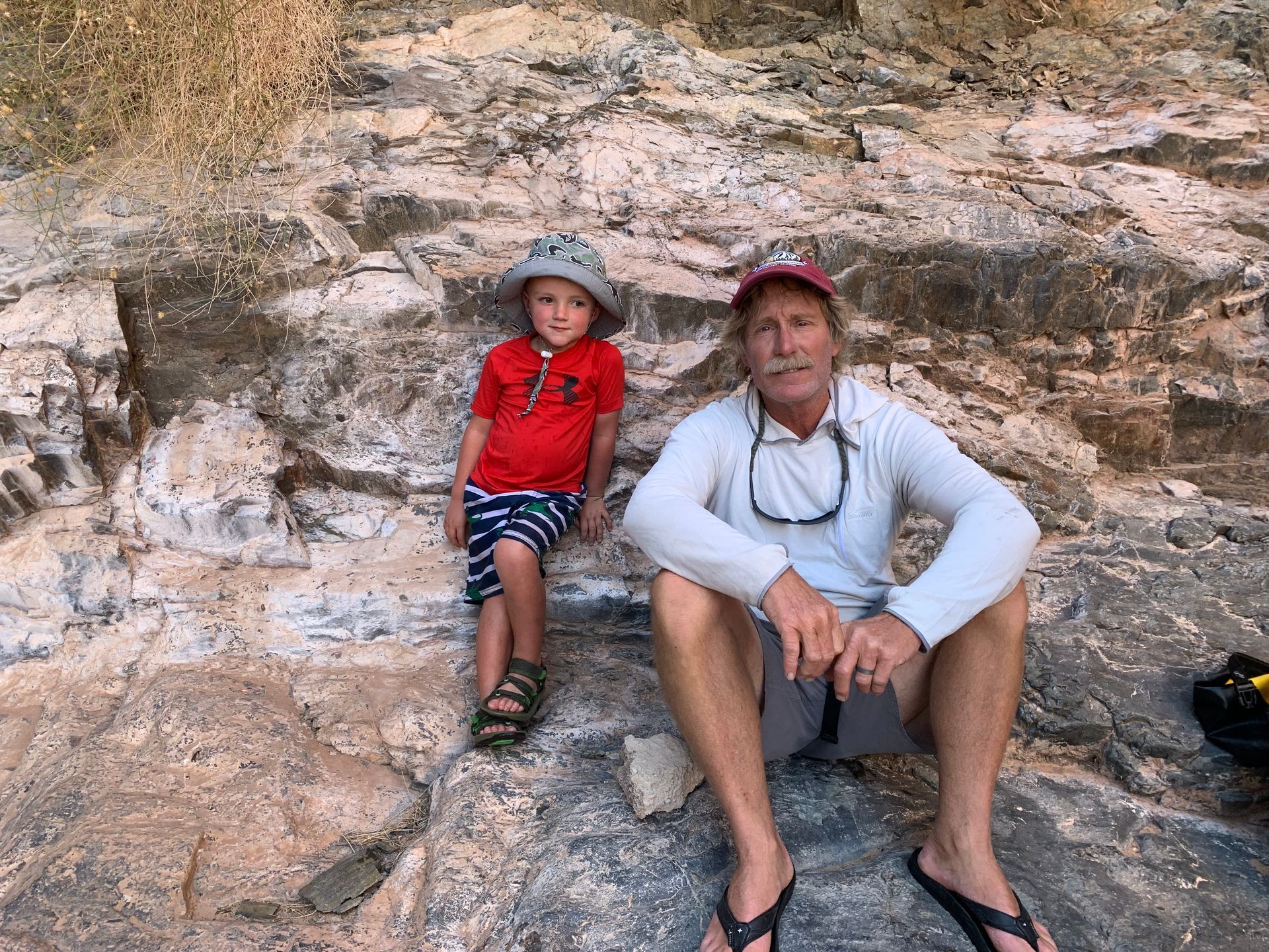 Man and child sitting against a rocky backdrop. Child wears red shirt, hat, sandals. 