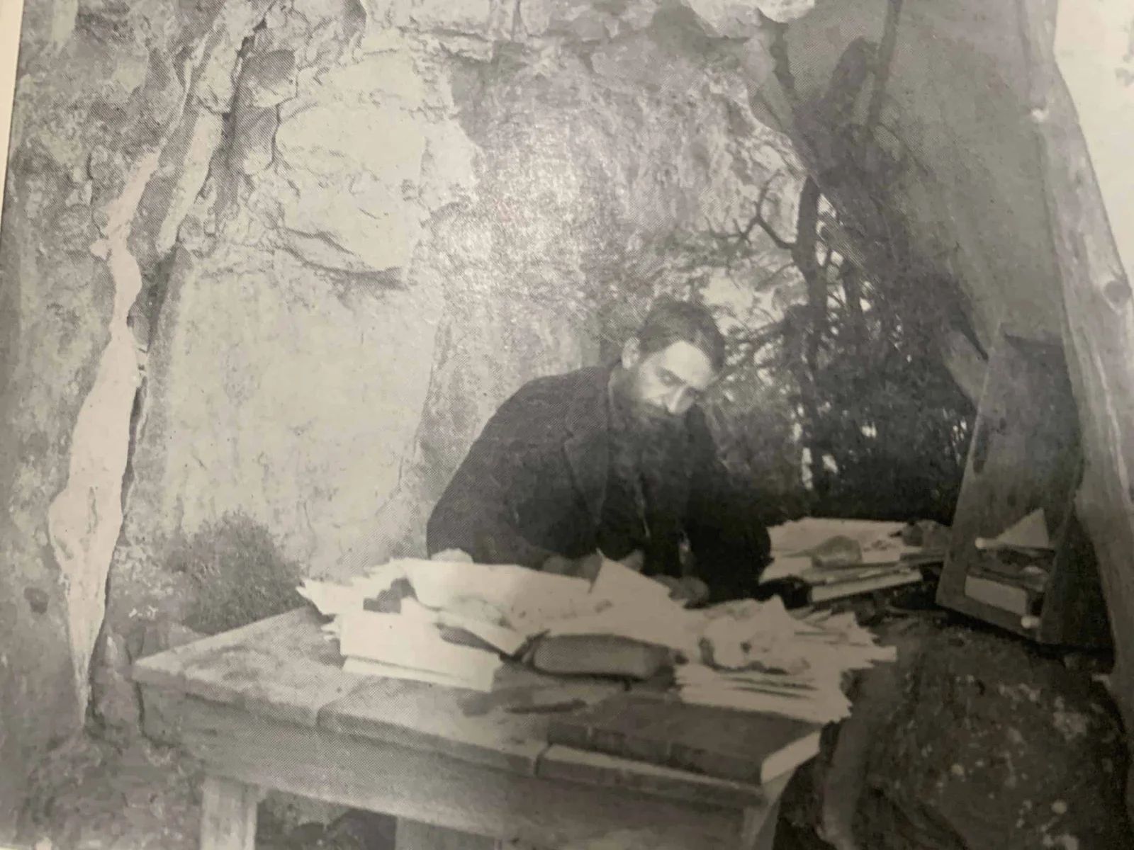 Man in dark robes at desk in a cave, surrounded by papers.