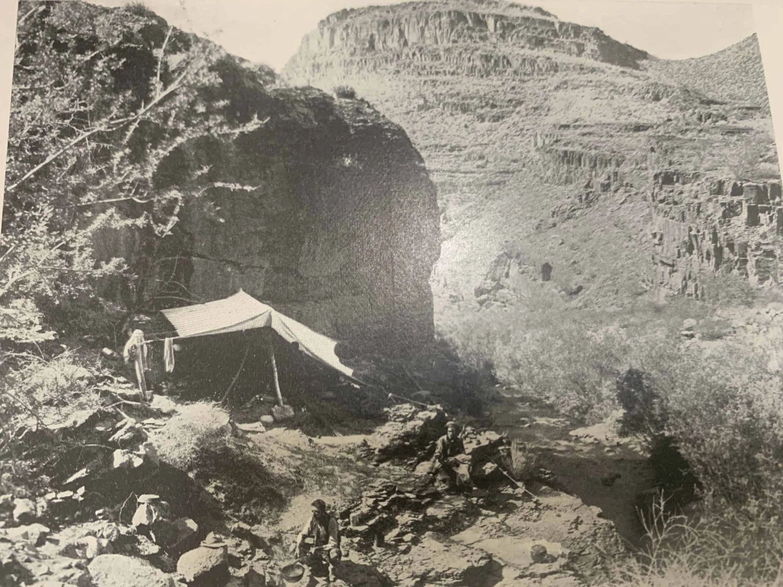 Tent set up near large rock formation in a canyon. Mountain in the background. Black and white.