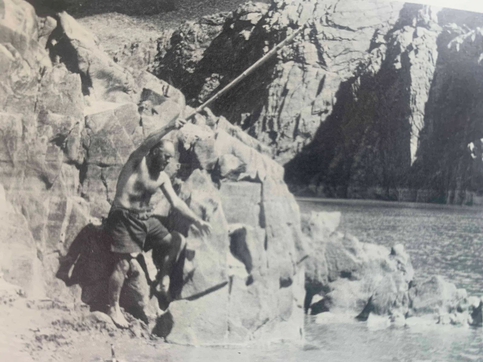 Man carving rock near water, using a long tool. Rocky cliffs and water in background.