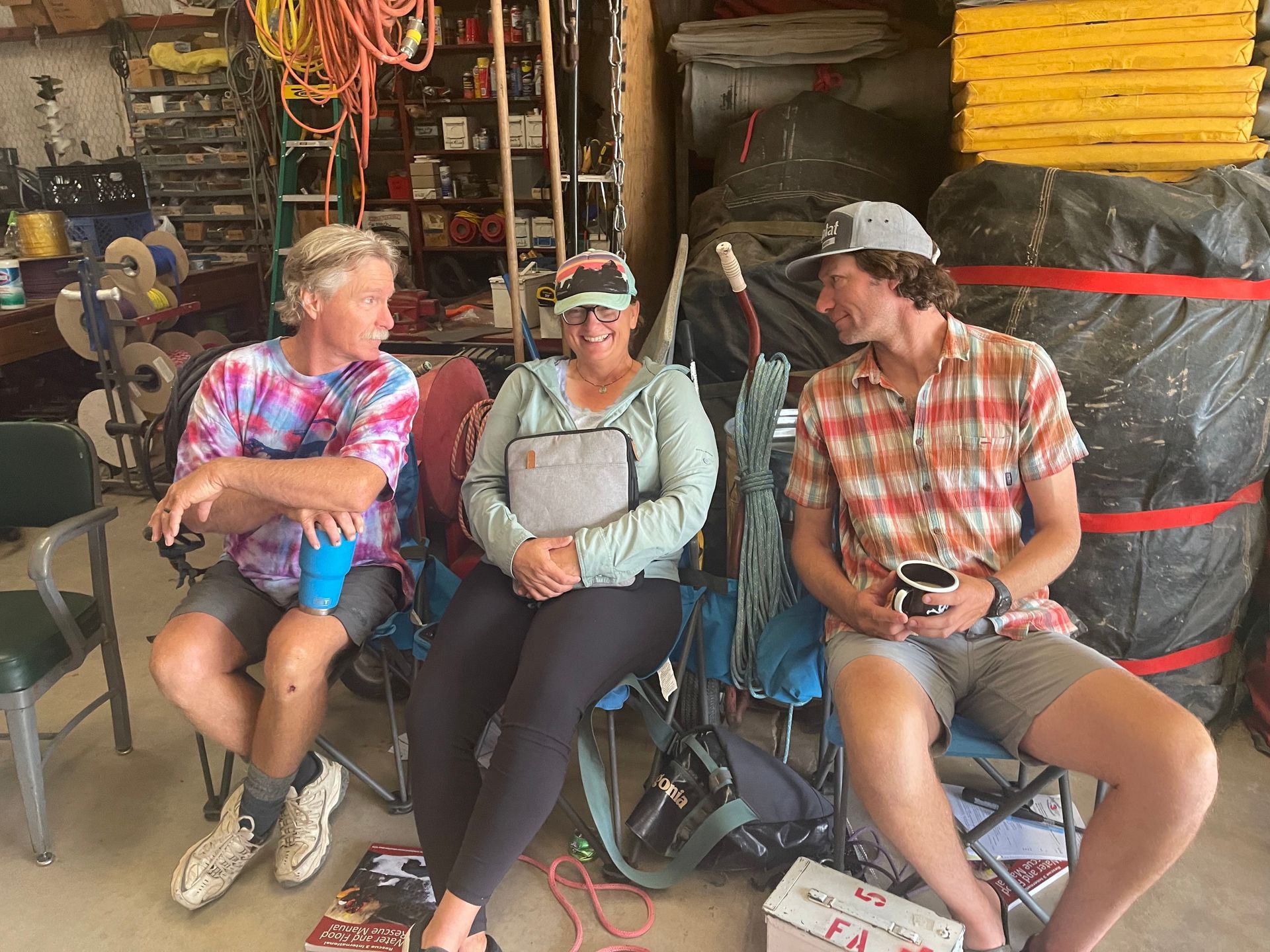 Three people seated, talking in a garage. One in tie-dye shirt, one with a laptop, the other with a cup.