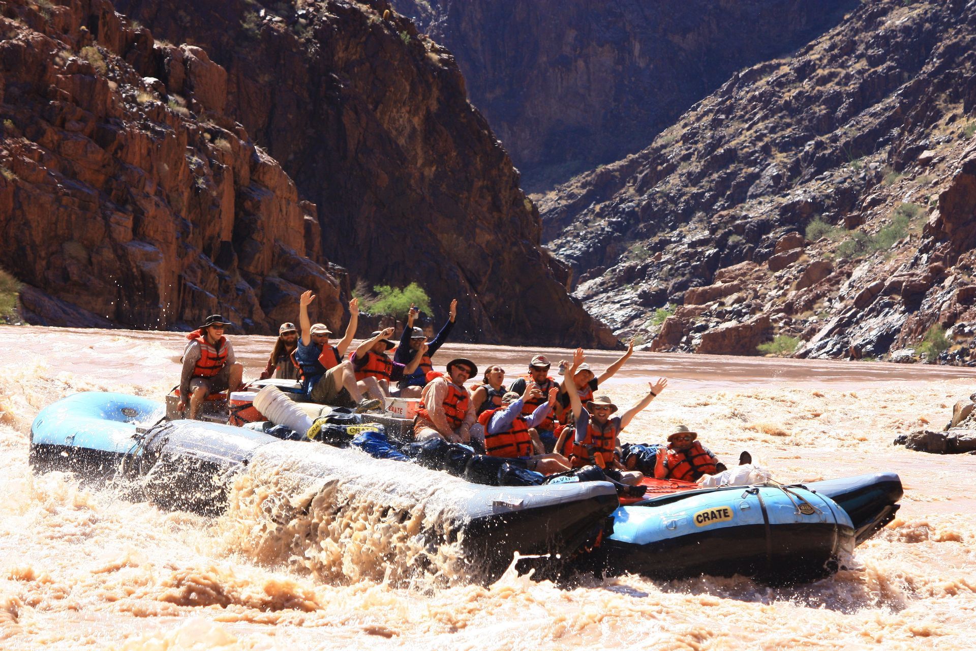 People in a raft on a rushing, brown river between canyon walls; some raise their arms.