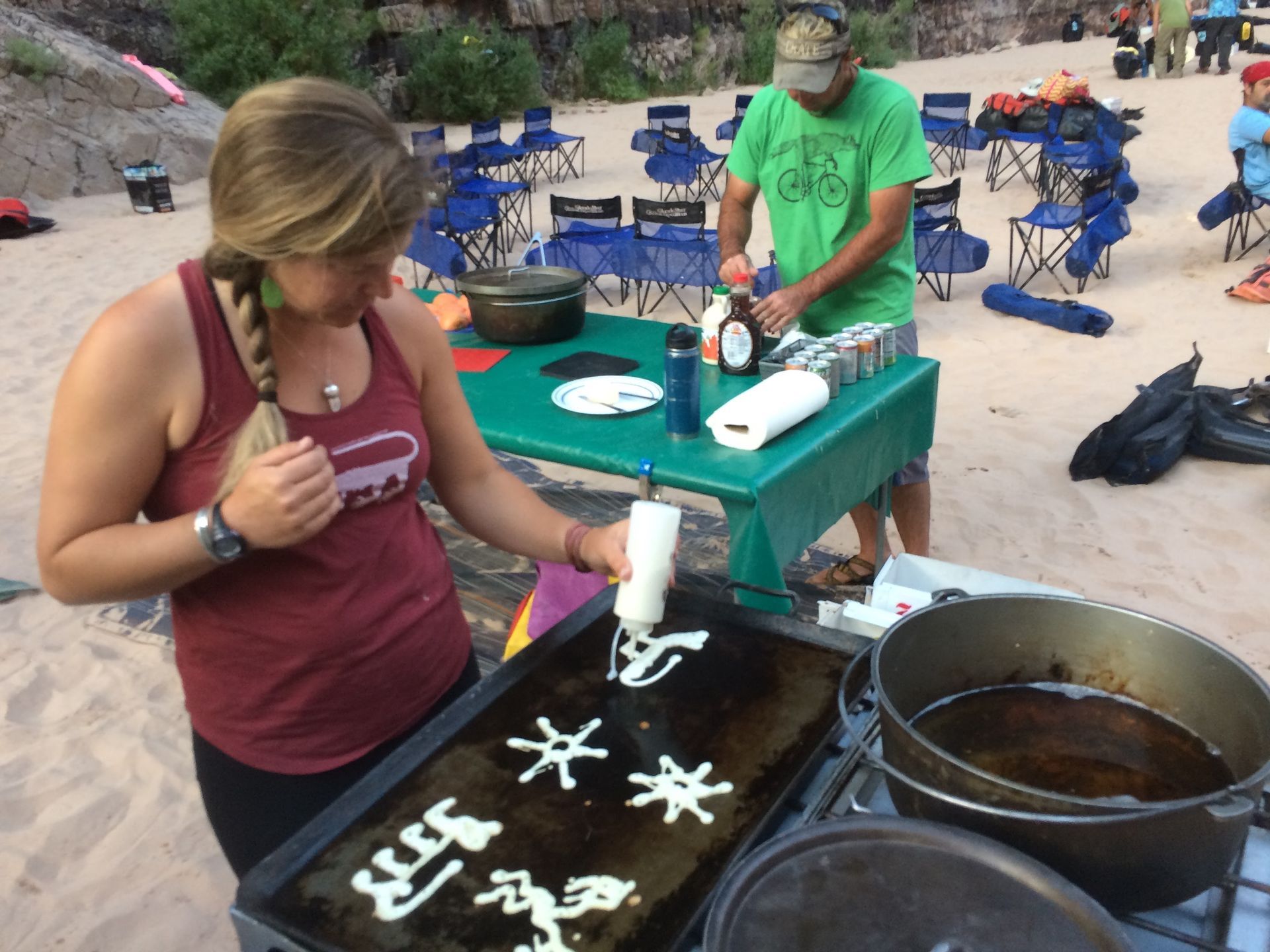 Woman squeezing batter onto griddle, making shapes. Man stands nearby. Outdoor cooking scene.