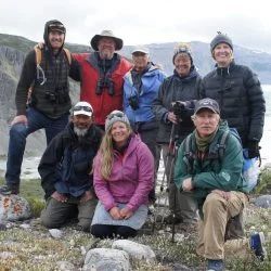 Group of hikers on a rocky landscape; some smiling, wearing jackets, hats, and carrying gear near a glacier.