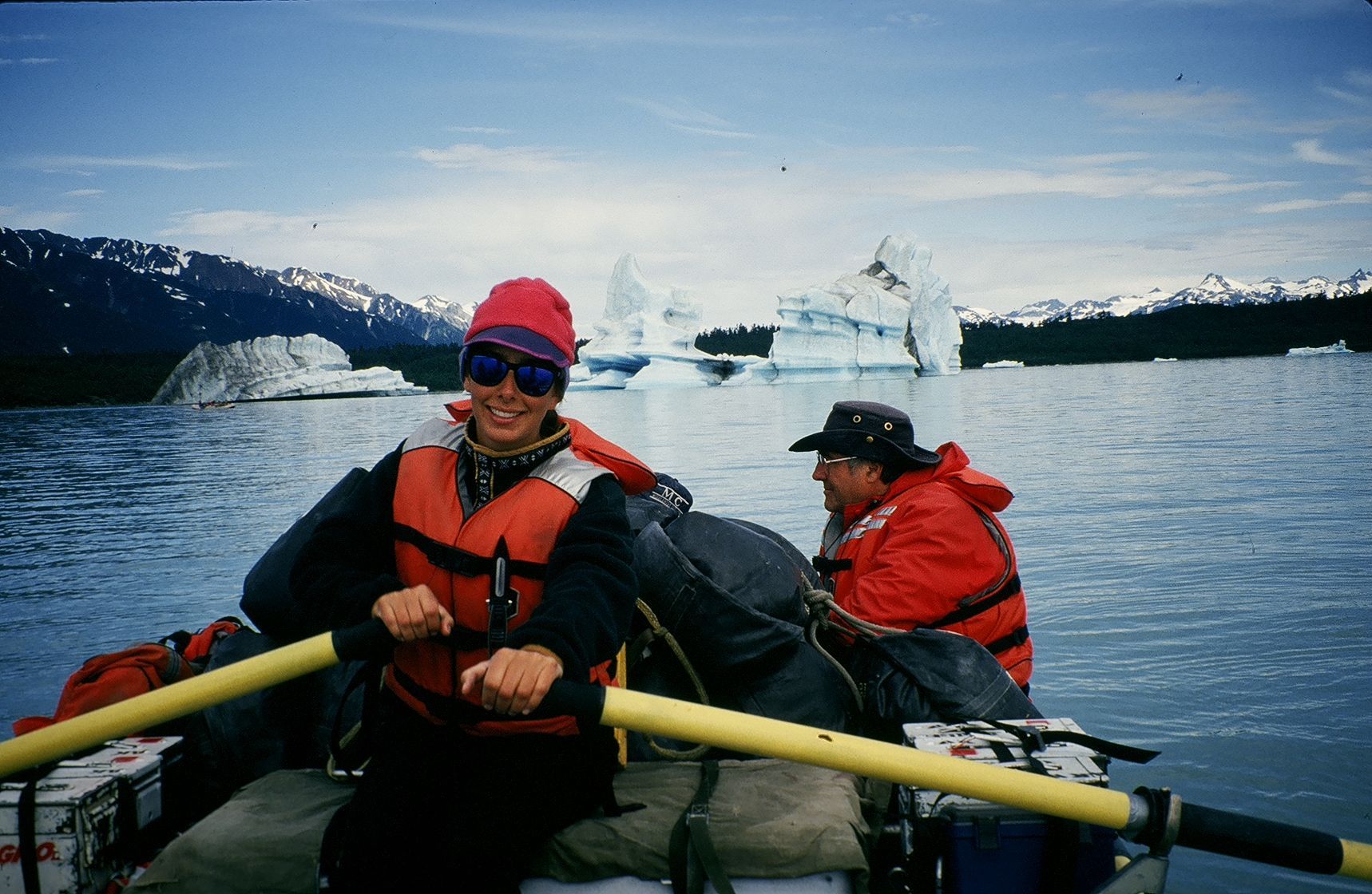 Two people rowing a raft on a lake, icebergs in the background. Woman in red hat and life vest smiles.