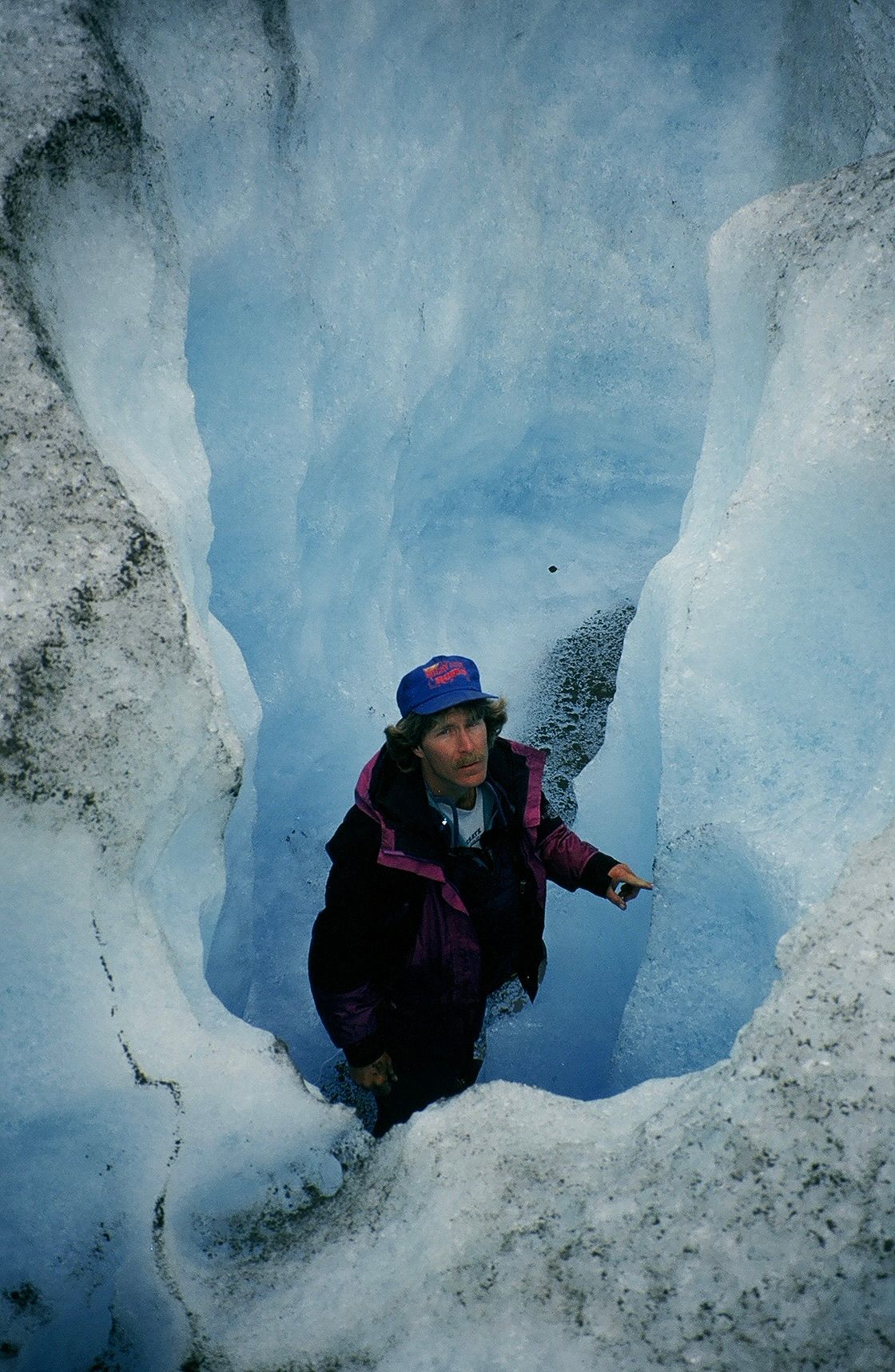 Person standing in a glacial crevasse, surrounded by blue ice. They wear a hat and jacket, pointing.
