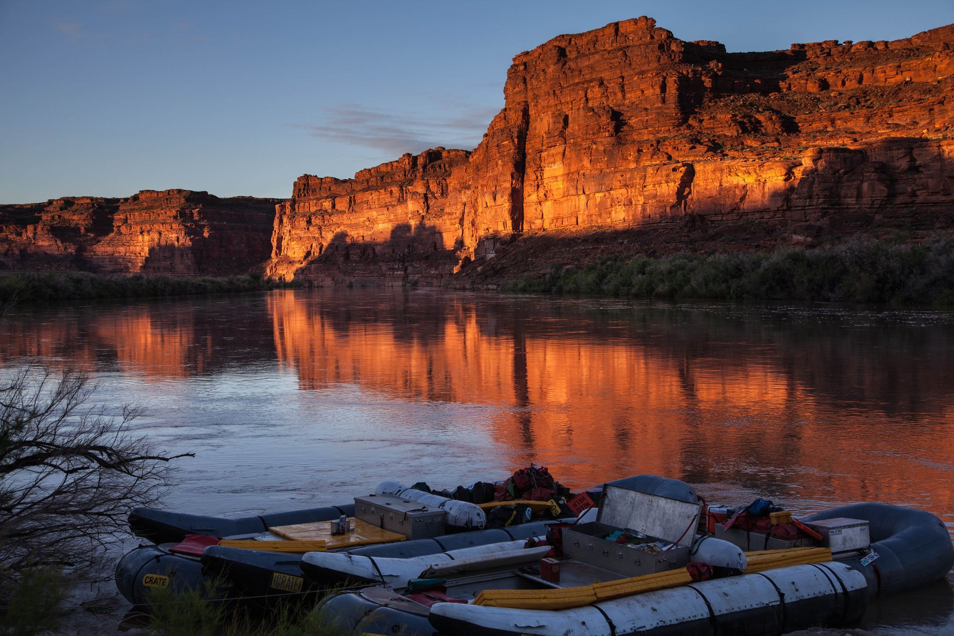 Rafts on a riverbank reflect the golden light of a towering canyon at sunset.