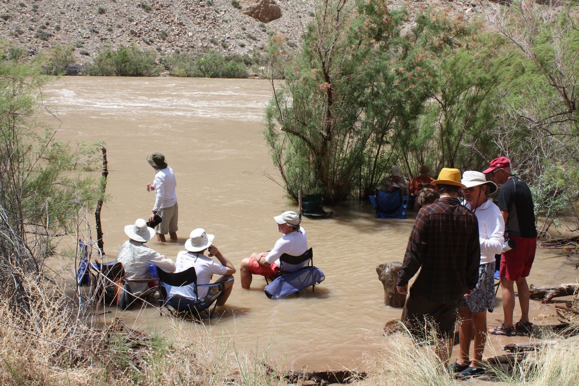People in hats and light clothing near a muddy river, preparing to possibly enter the water.