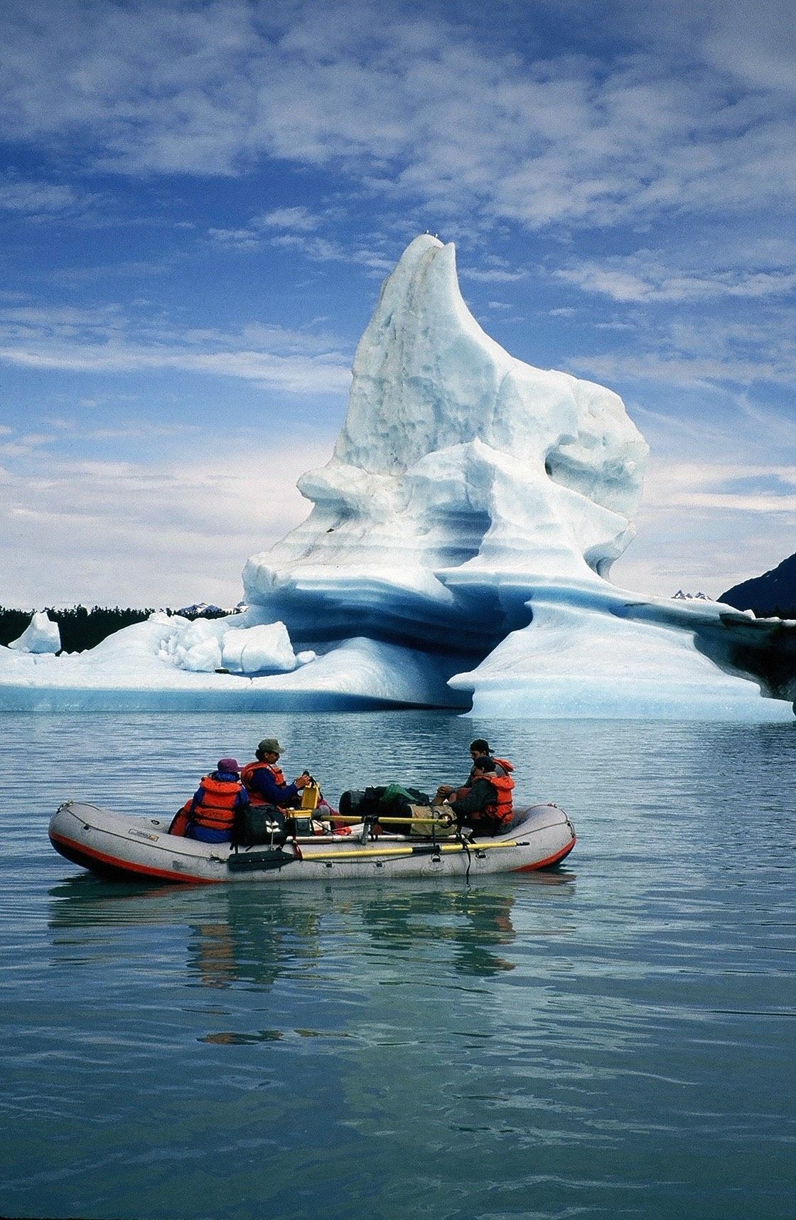 People in a raft near a large iceberg, blue water and sky.