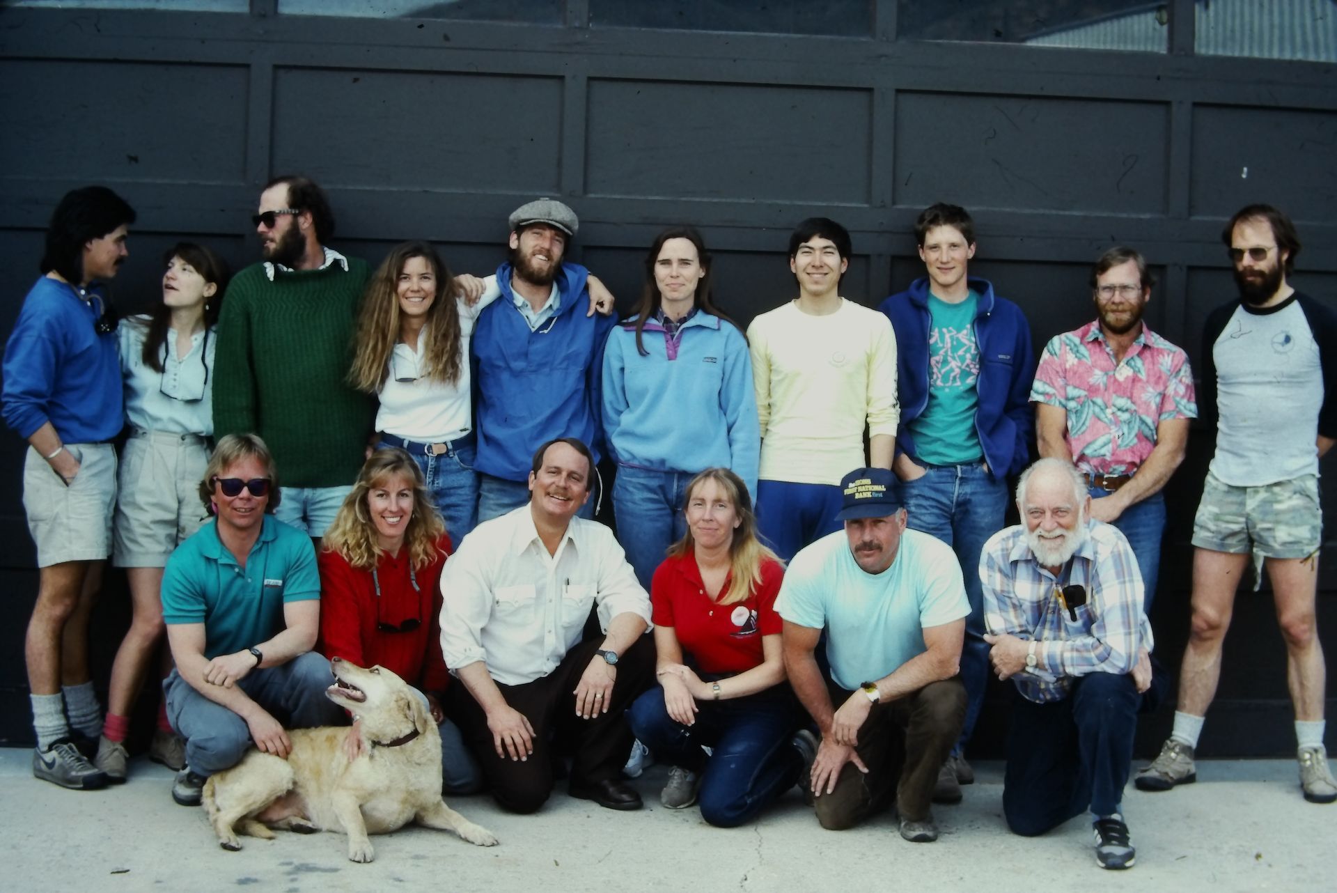 Group of people posing in front of a dark garage door. Some are standing, others kneeling, with a dog.