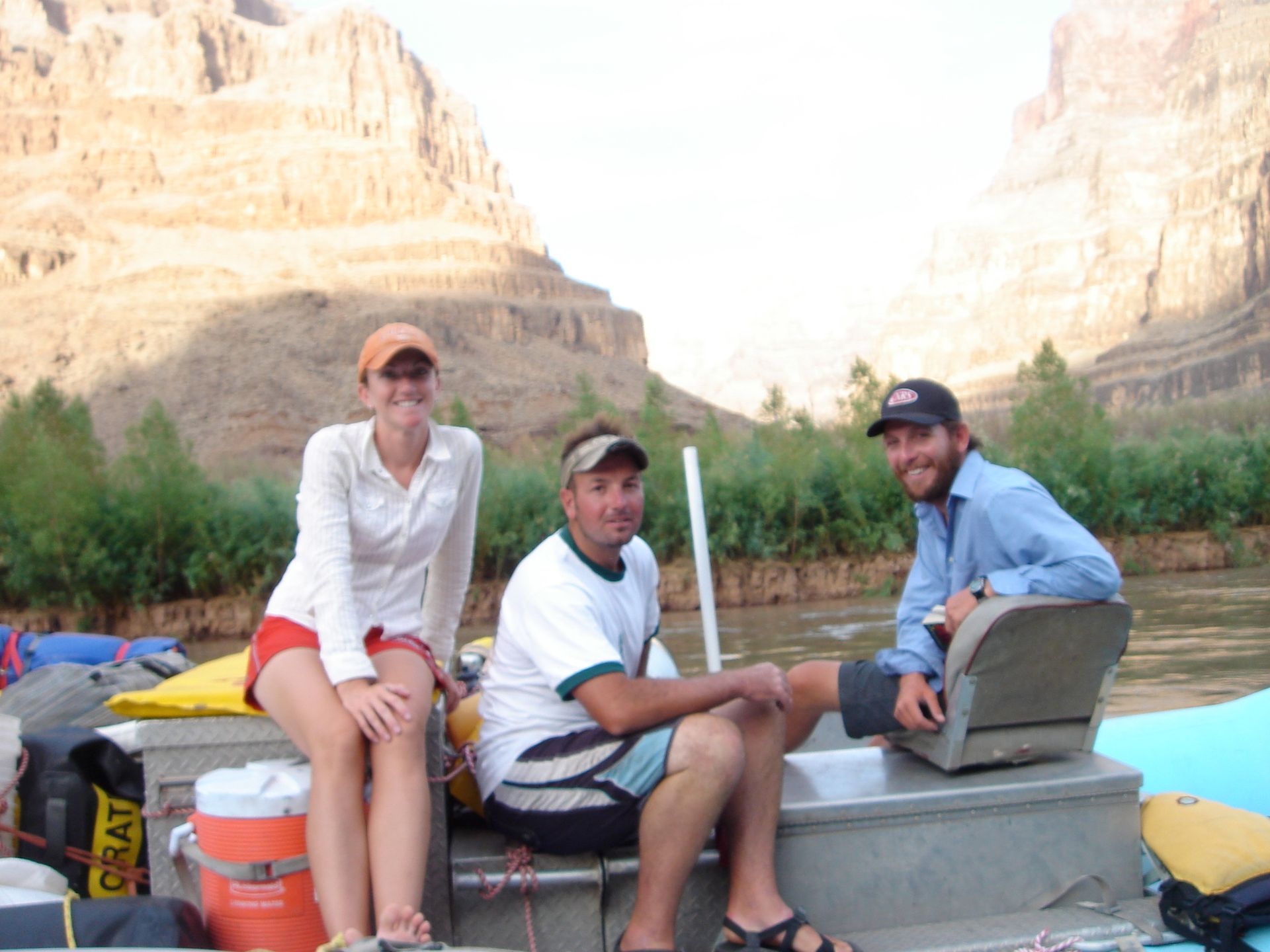 Three people on a boat in a canyon. Woman in hat, shorts. Men in hats and shorts. Canyon walls in background.