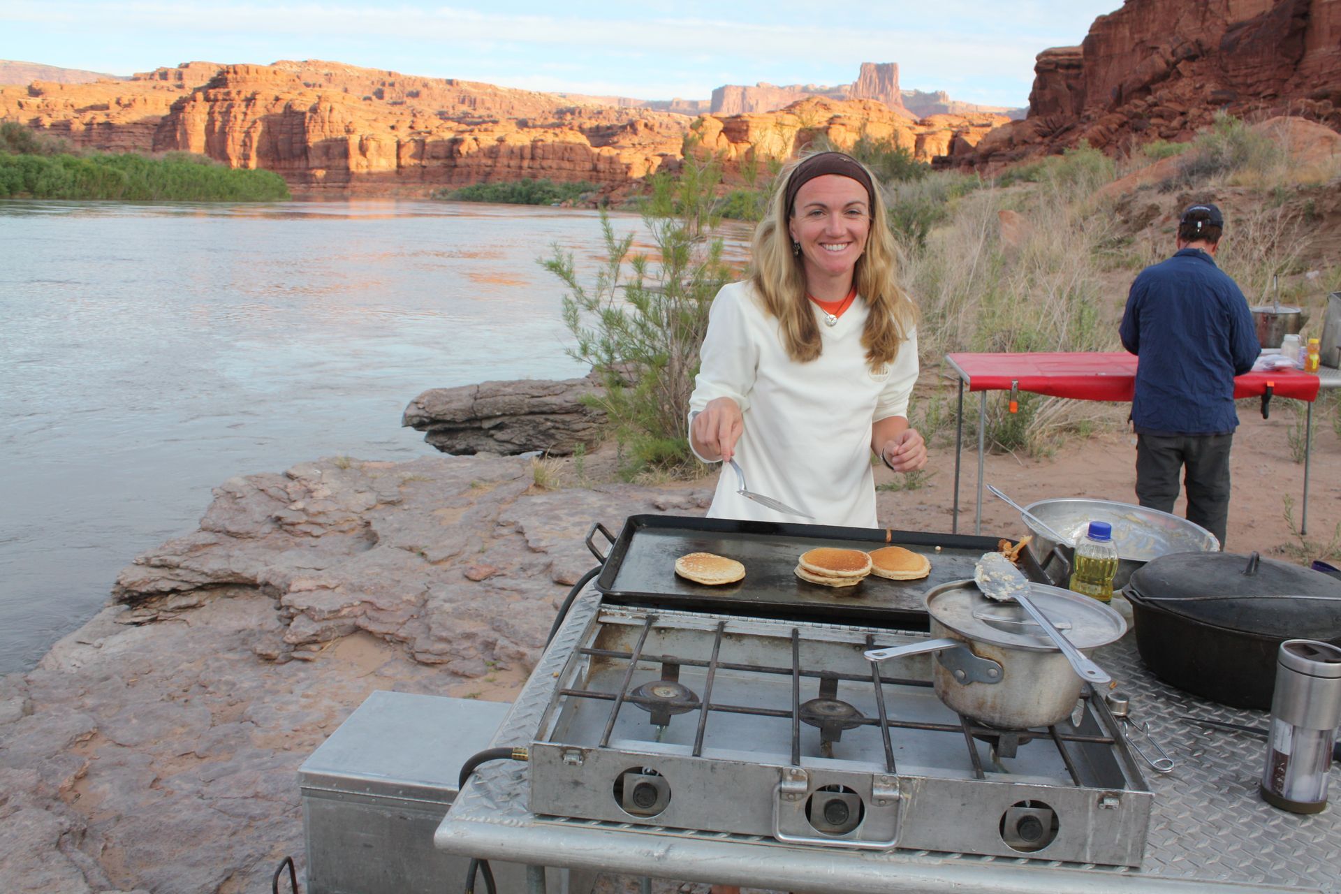 Woman cooking pancakes by a river, with red rock formations in the background. Man standing nearby.