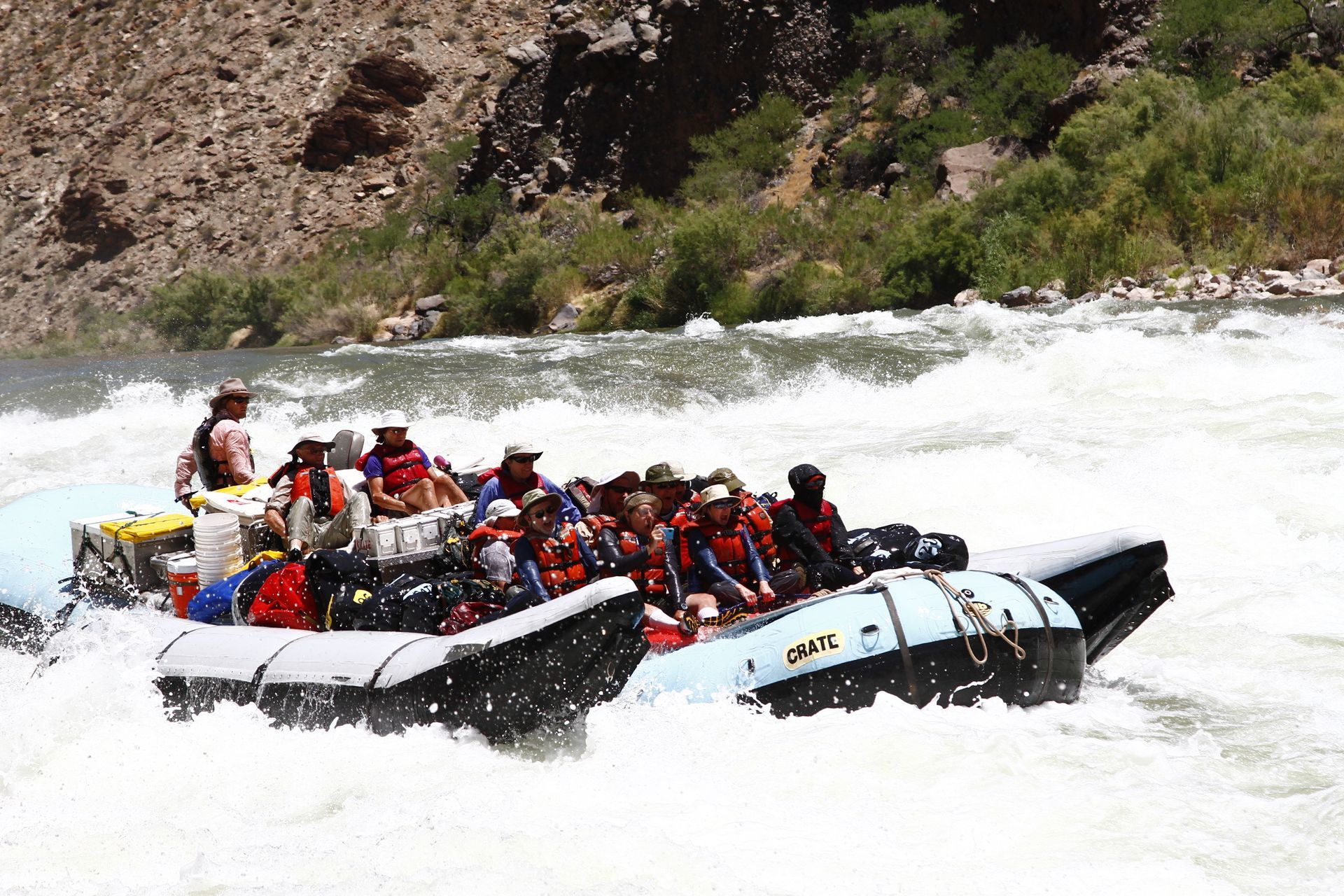 Raft of people navigating a whitewater rapid. Rocky canyon walls in background.