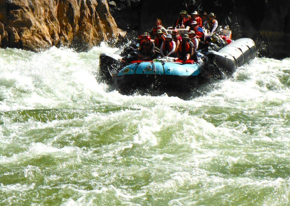 Raft with people navigating whitewater rapids; water splashes, sunny setting.