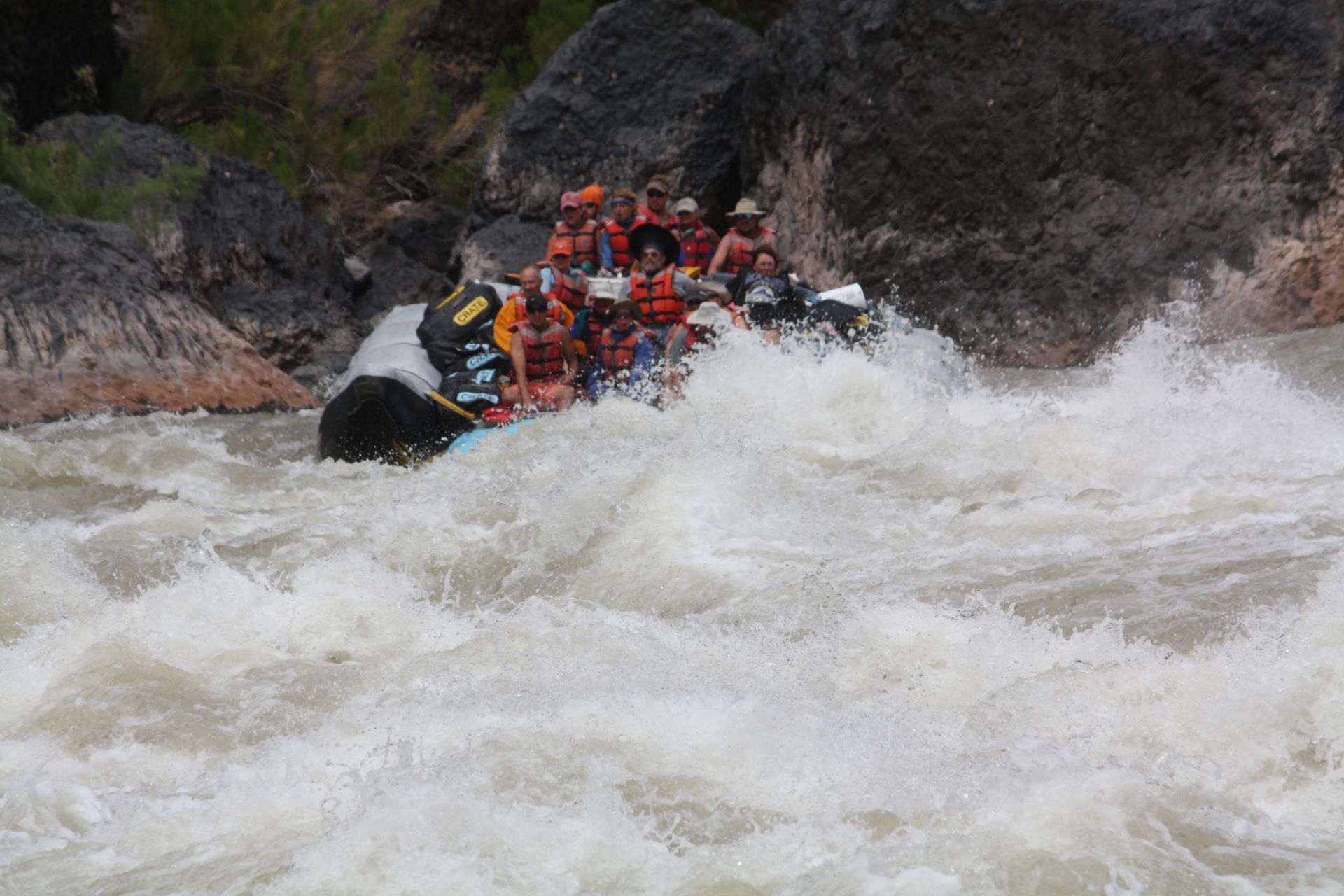 People on a raft navigate a whitewater rapid. They wear life vests, and the water is turbulent.