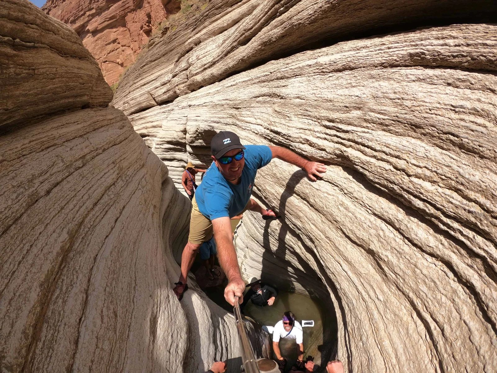 Man climbing a narrow rock canyon, holding a selfie stick. Others below. Beige canyon walls, blue shirt.