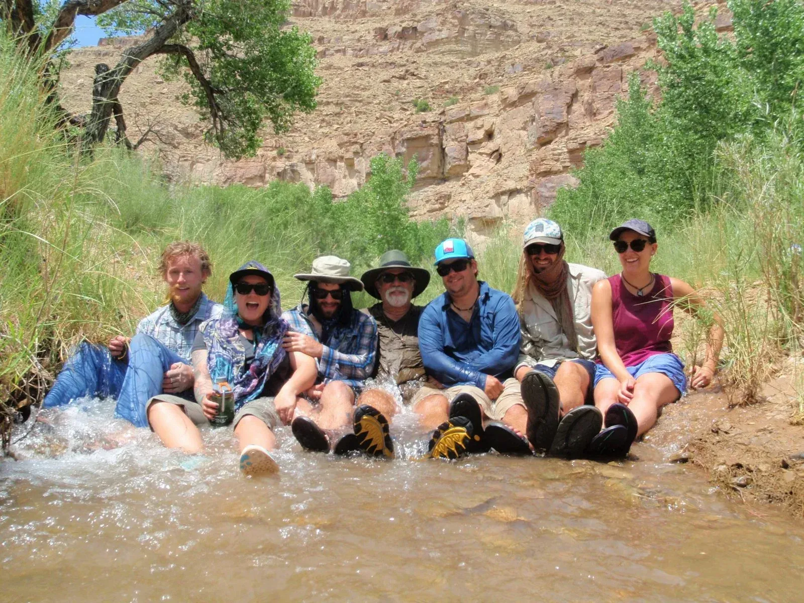 Group of people sitting in a stream; mountain backdrop, sunny day, water splashing.