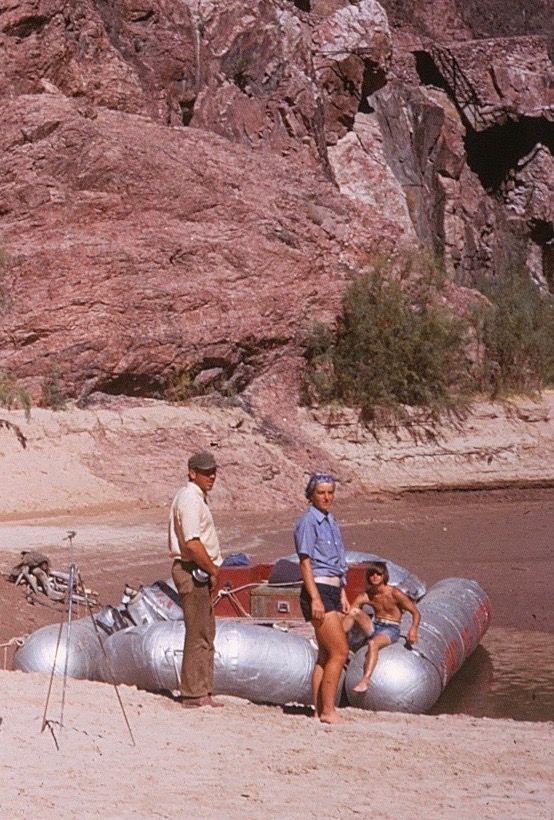 Two people stand near a silver raft on a sandy shore next to a red rock cliff.