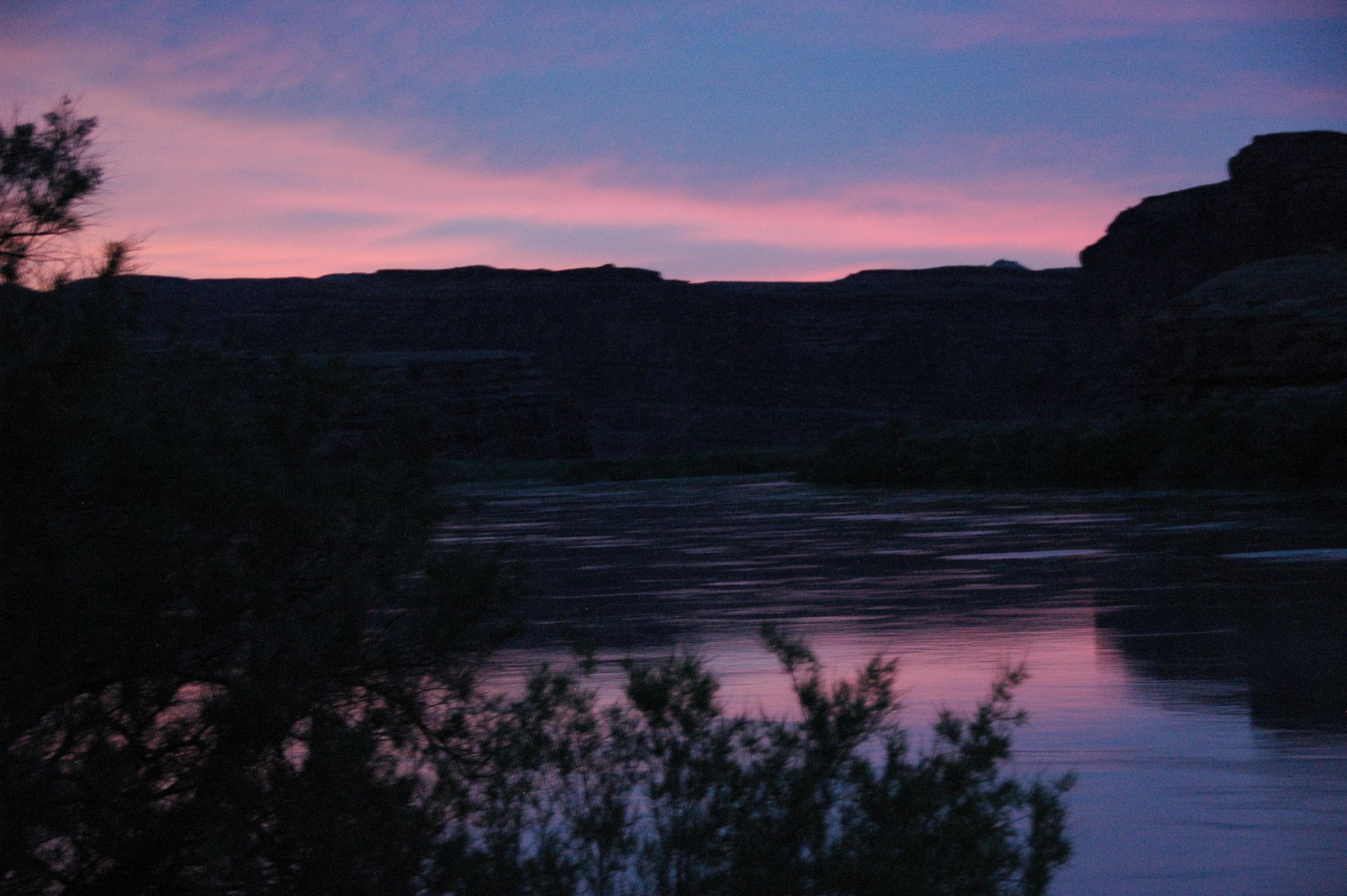 Pink and purple sunset reflects on a calm river; dark silhouette of land and vegetation.