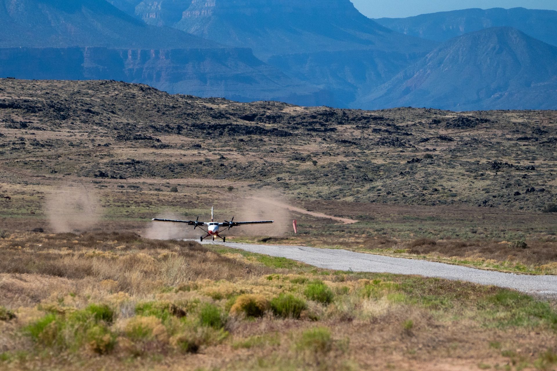 Airplane taking off from a dirt airstrip, kicking up dust with mountainous backdrop.
