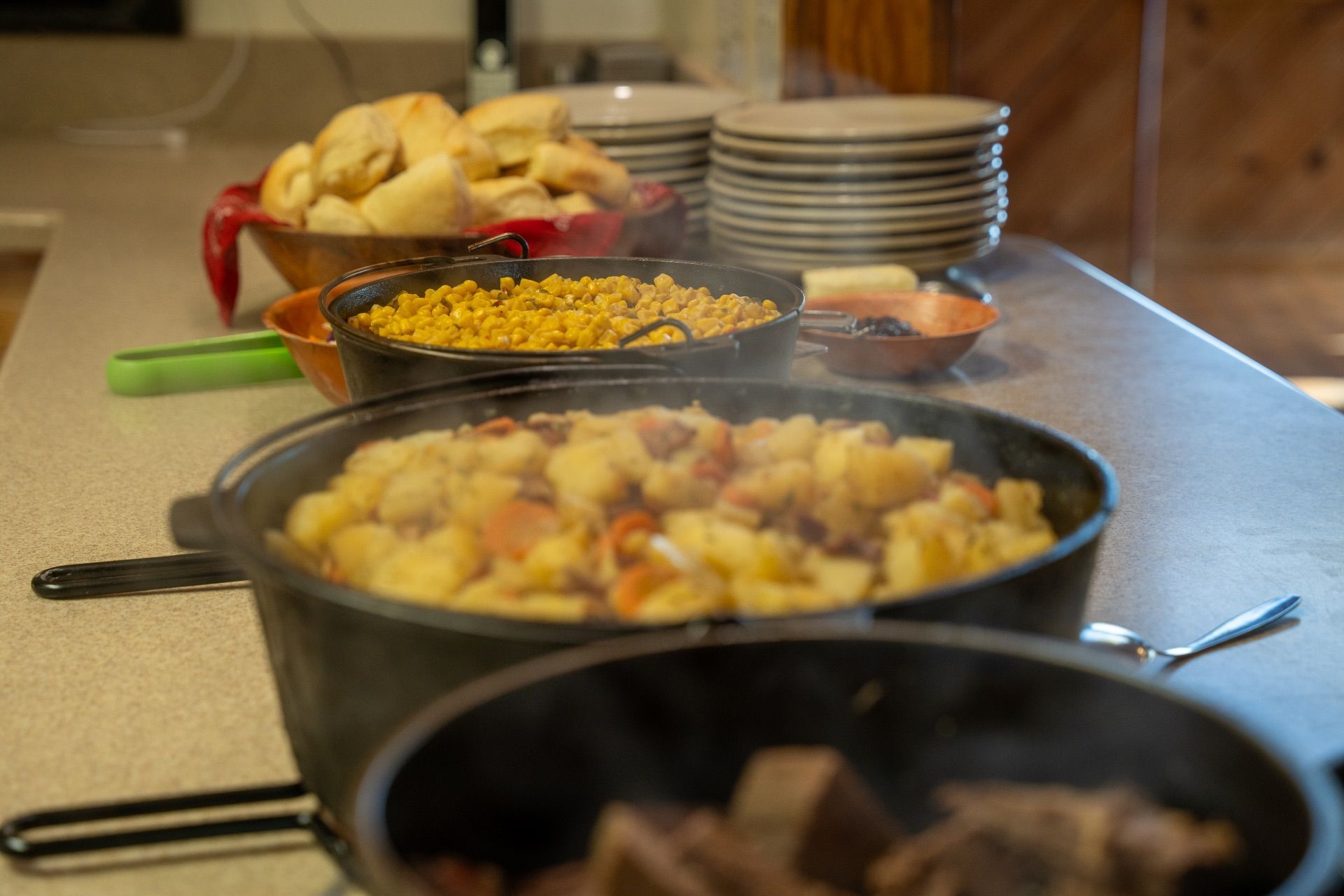 Buffet with cast iron pots of food, rolls, and plates on a counter.