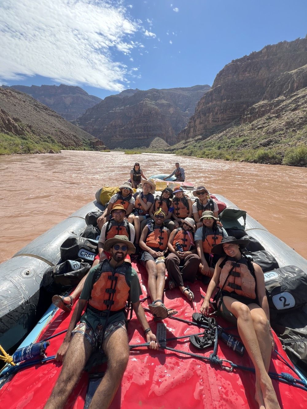 People in life vests on a raft in a red river, canyons in the background. Sunny day.
