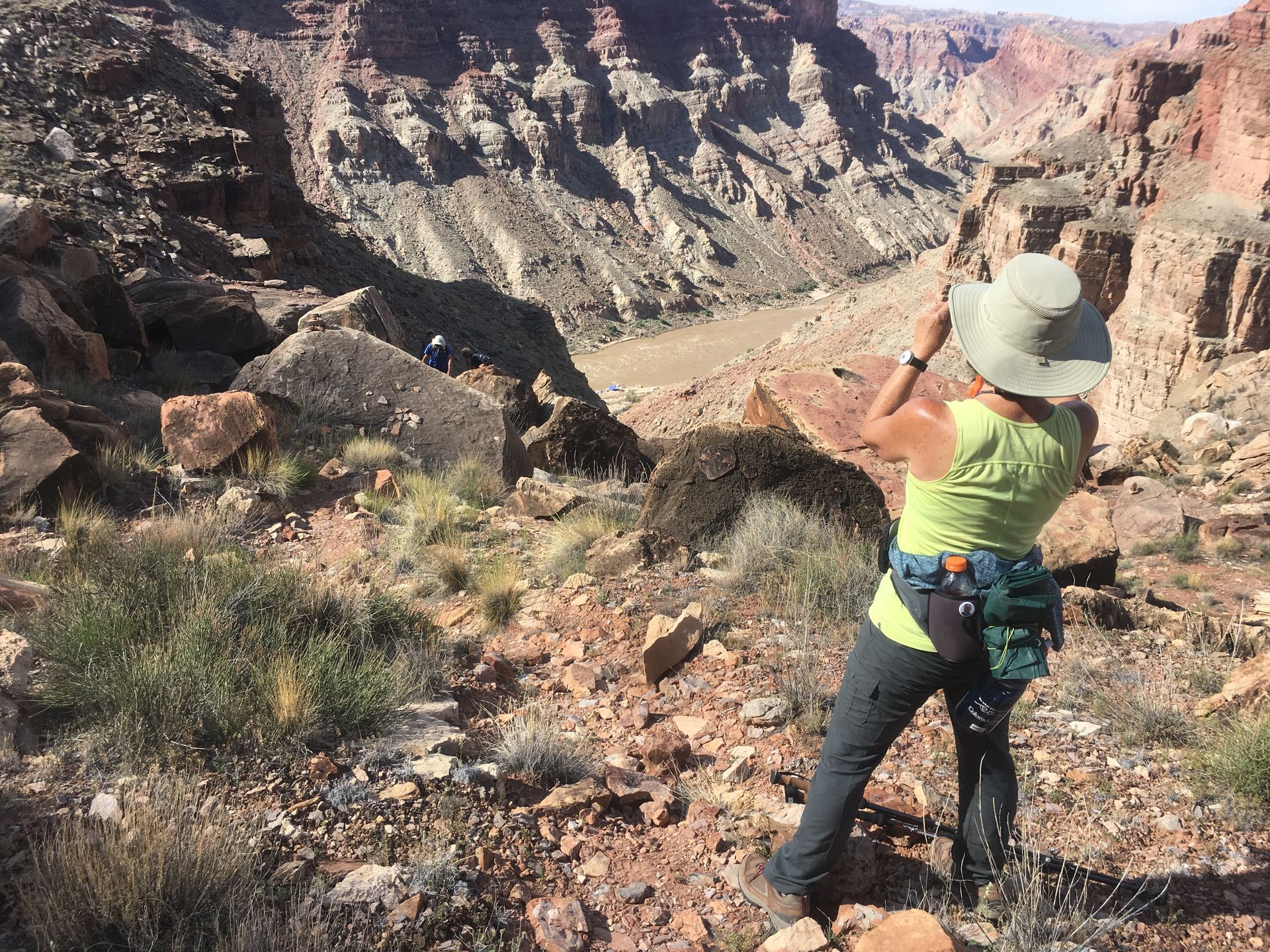 Woman in a hat taking a photo of a canyon. Rocky terrain, light and dark tan canyon walls.