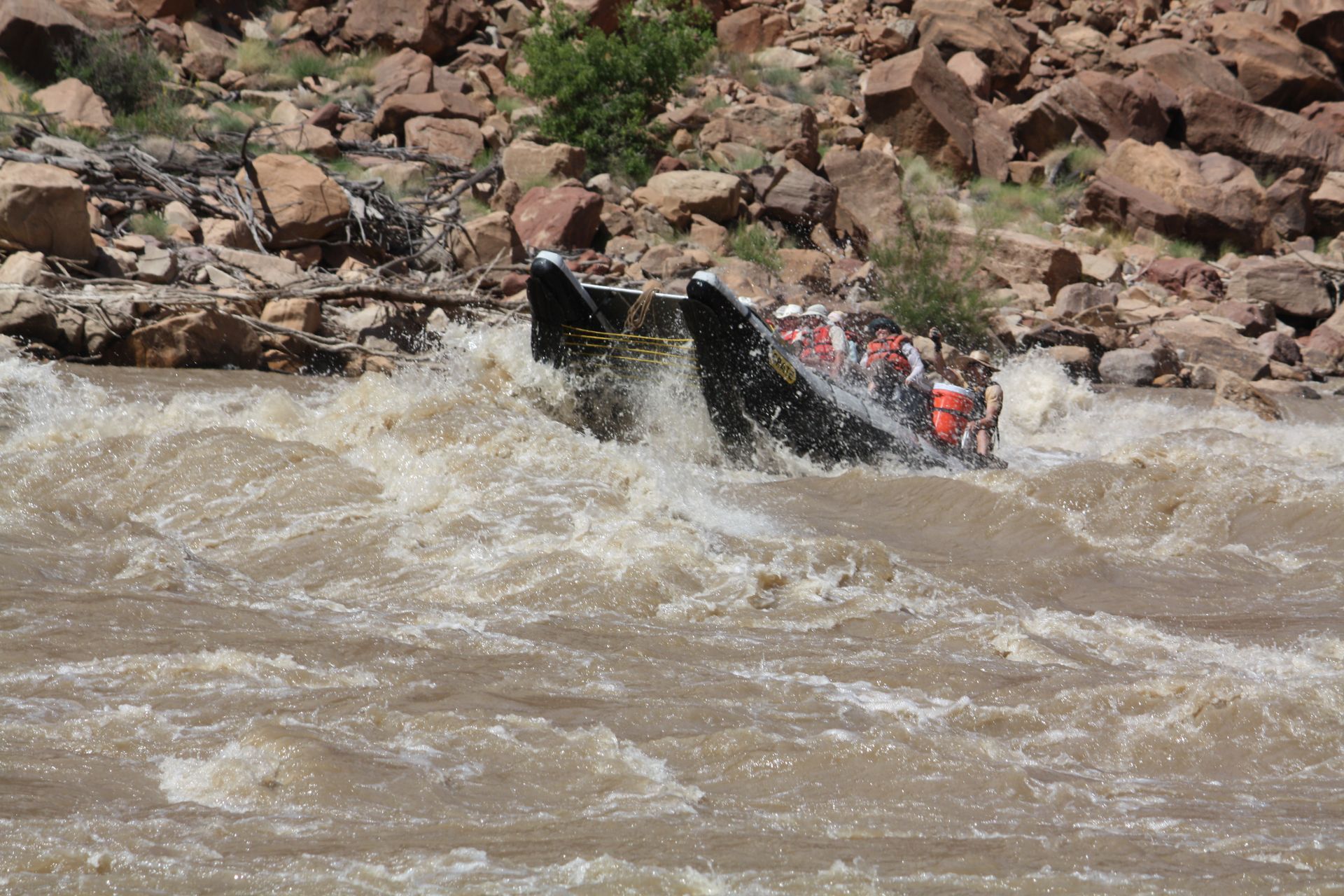 Whitewater rafting boat navigating rapids in a brown river, rocky banks in the background.