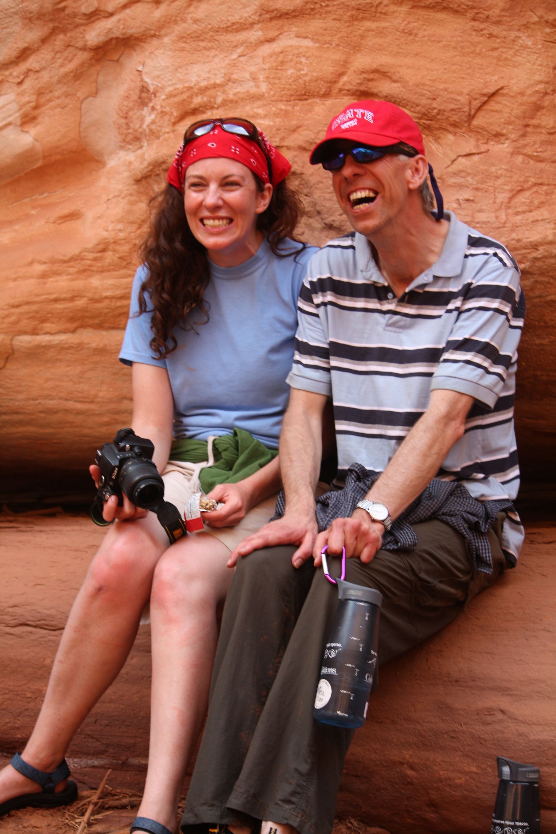 Woman and man laughing, sitting outdoors against a red rock background. Woman holds camera. Man wears a red cap.