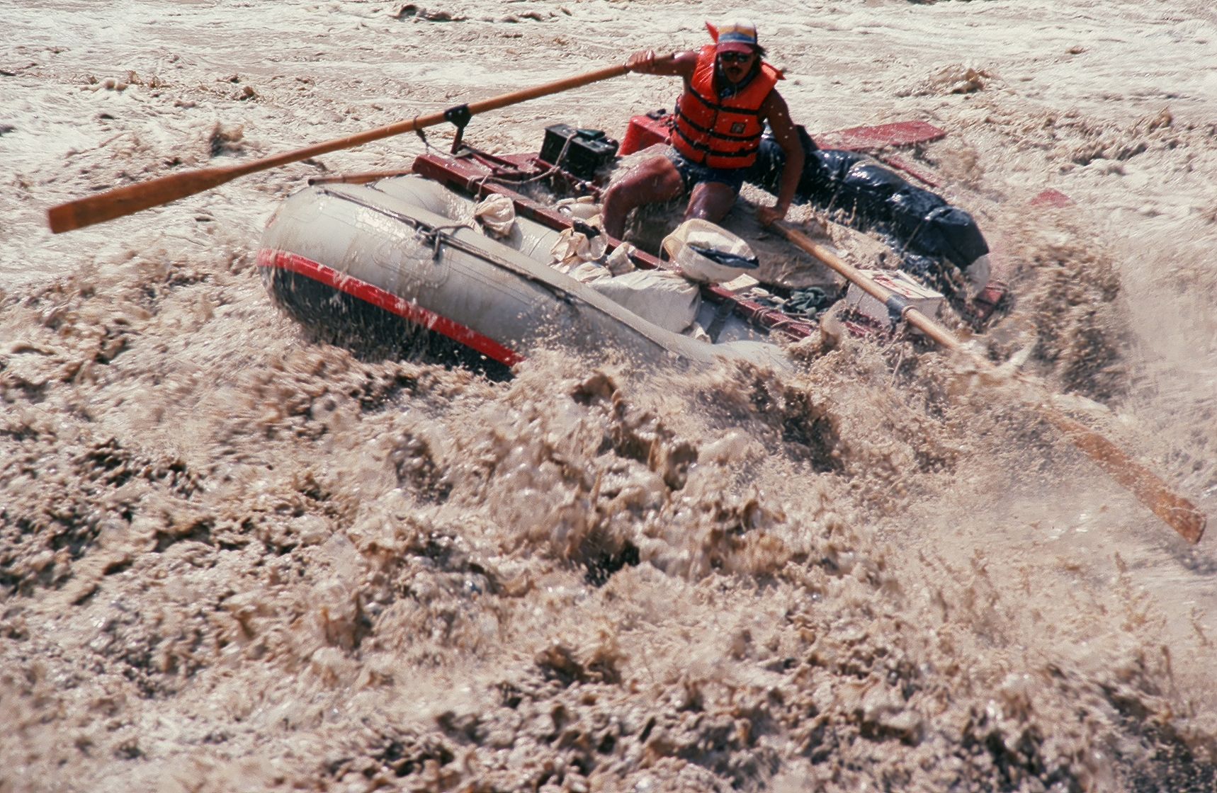 Man in orange life vest rows a raft through turbulent, brown water.