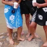 Three people wearing sarongs, holding mugs, standing on a rock near water.