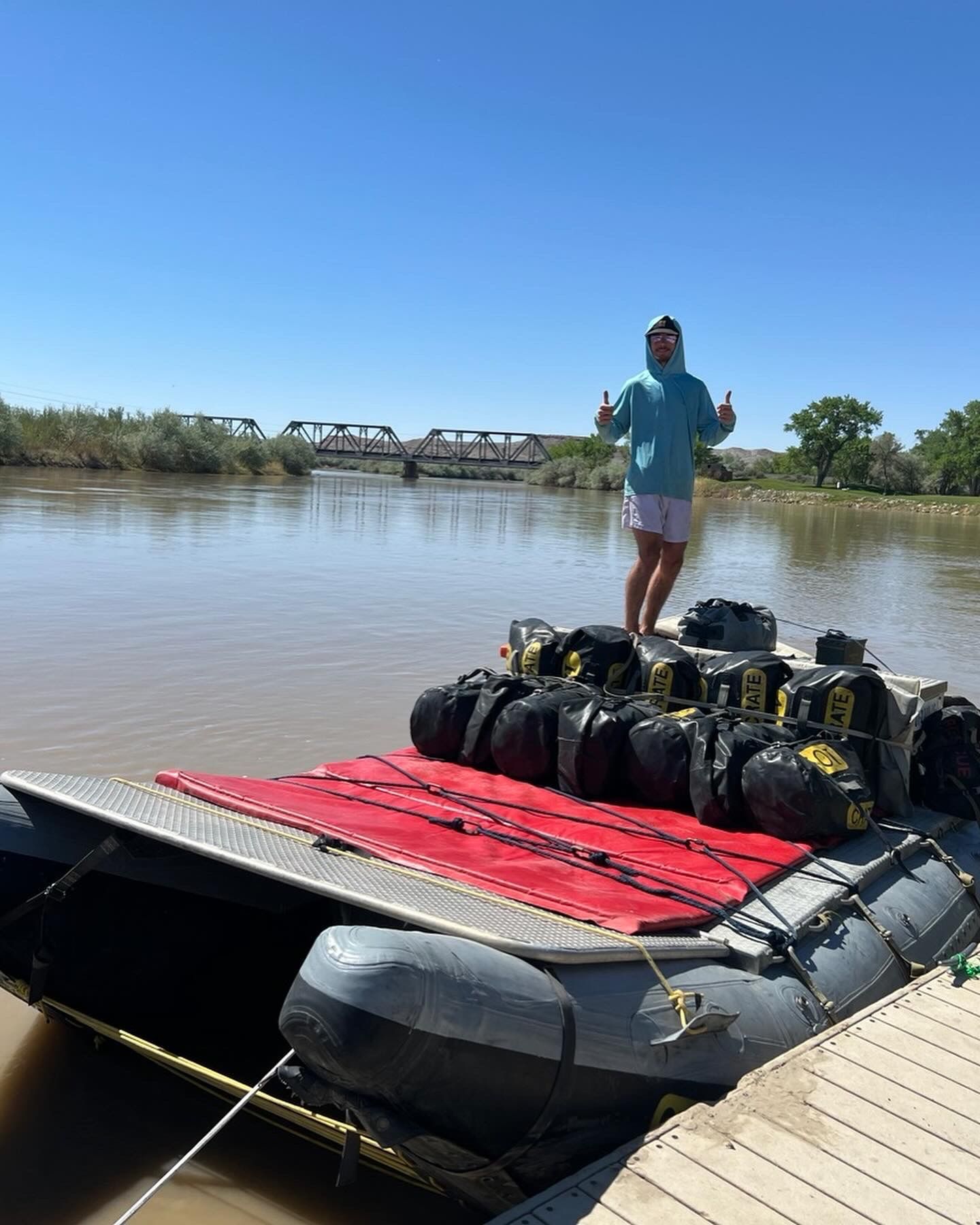 Person on a raft filled with gear, giving thumbs up, on a river near a bridge.