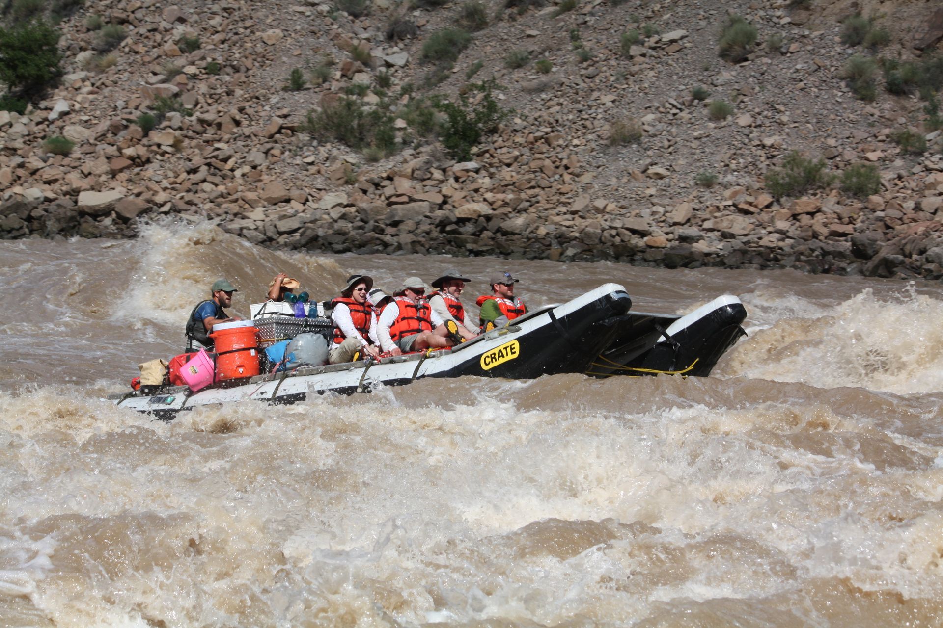 Rafting on a brown river, people in life vests navigate rapids near a rocky shore.