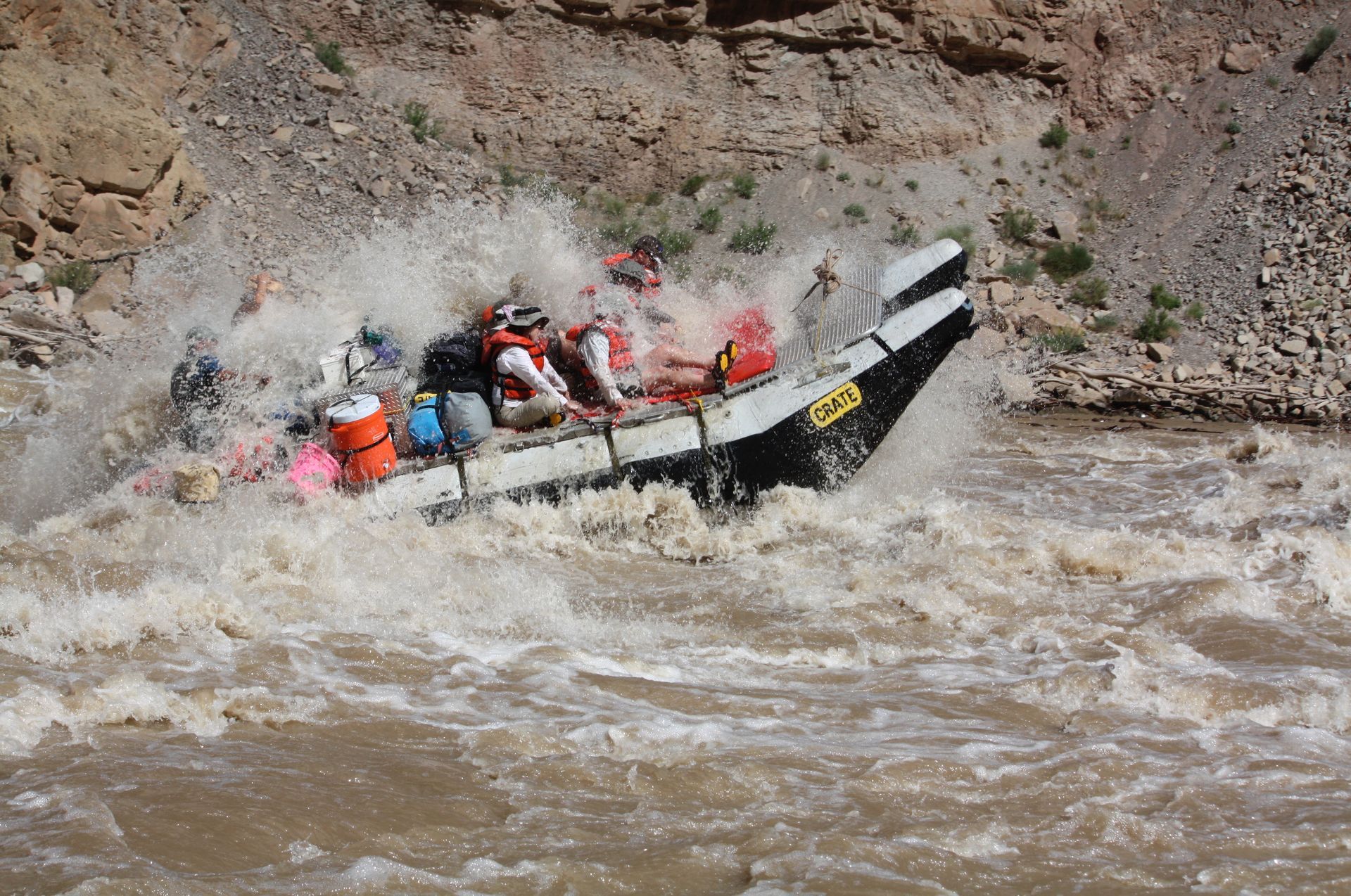 Two people on a raft in a canyon river, wearing life vests and hats.