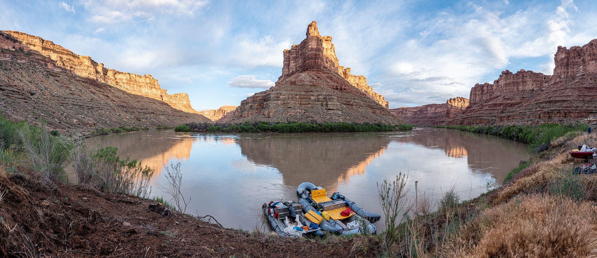 Panorama of a river with a rocky butte and canyon walls, and two rafts are moored on the bank.