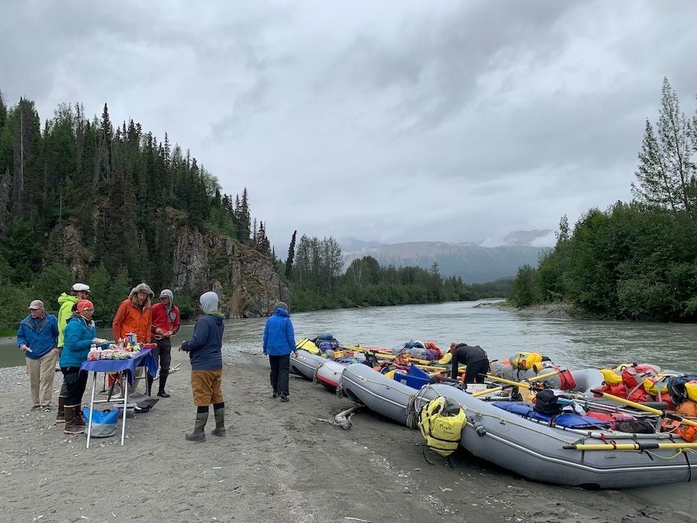 People preparing rafts on a riverbank with gear. Mountains and trees in the background under a cloudy sky.