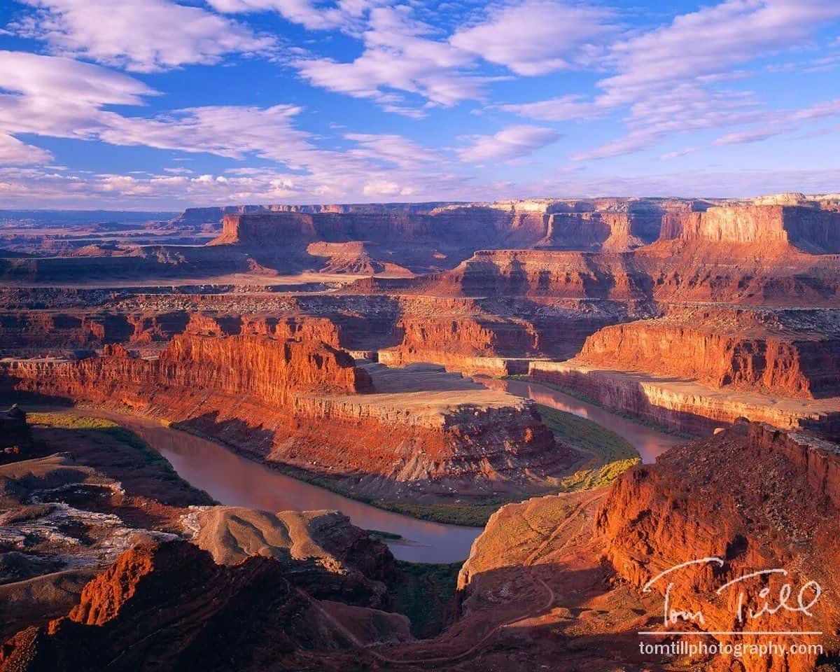 Red rock canyon landscape with a river winding through it under a partly cloudy blue sky.