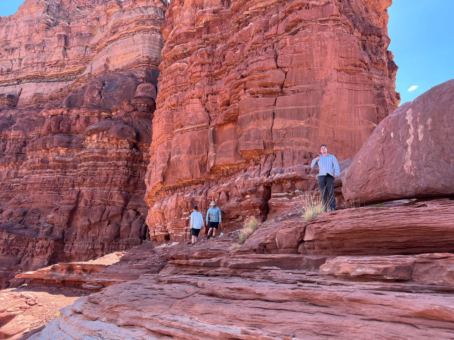 Three people hike near a towering red rock formation under a blue sky.