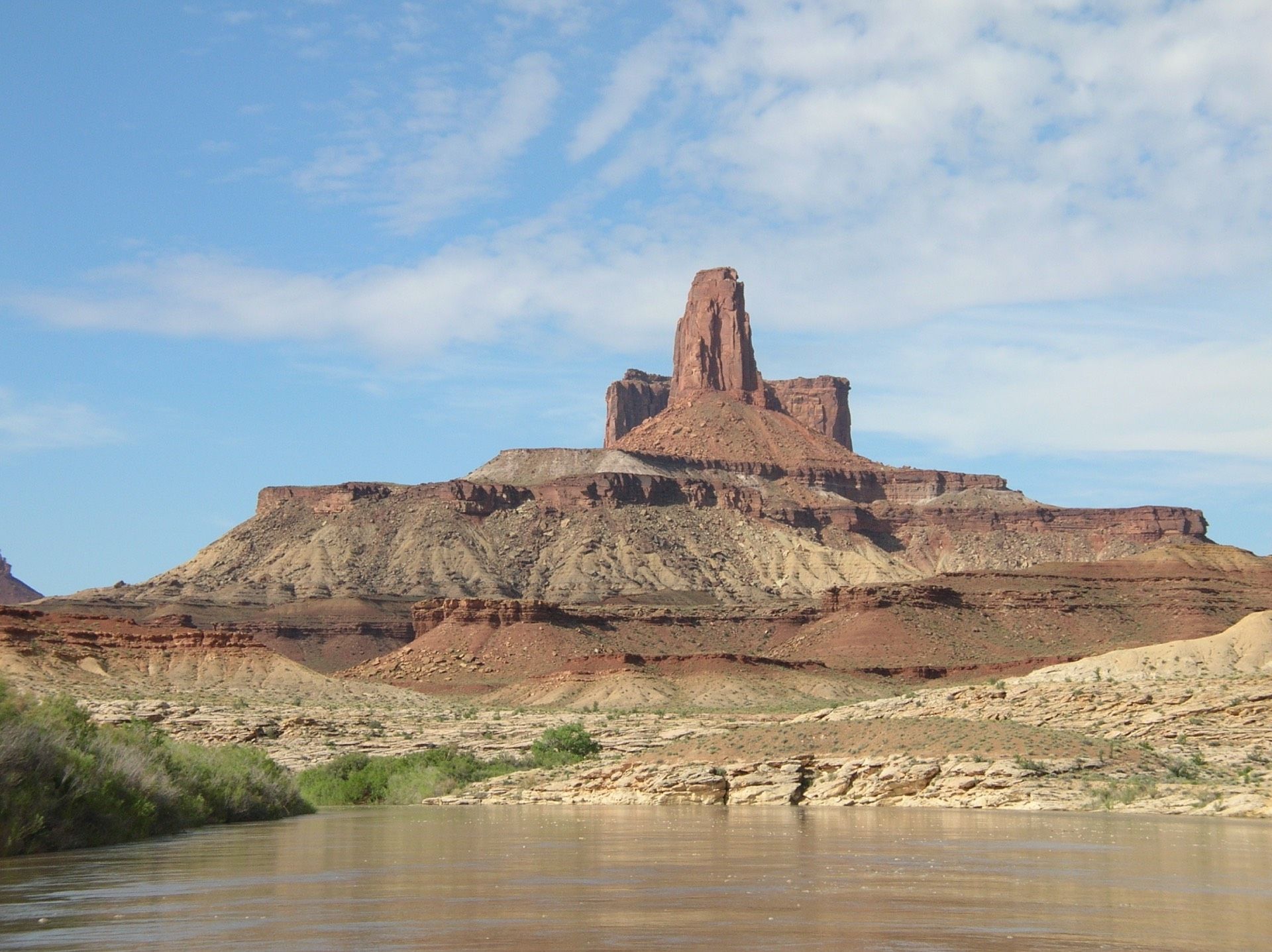 Red rock butte with layered cliffs, desert vegetation, and muddy river under a blue sky.