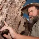 Man in safari hat points at rock face, appears surprised, outdoors.