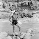 Woman standing on rocks near river, hands on hips, wearing shorts, top, and hat, against rock wall.