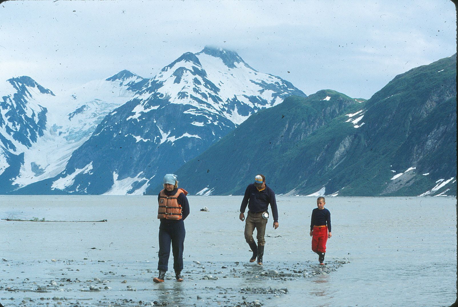 Three people walking on muddy shore in front of snow-capped mountains and a body of water.