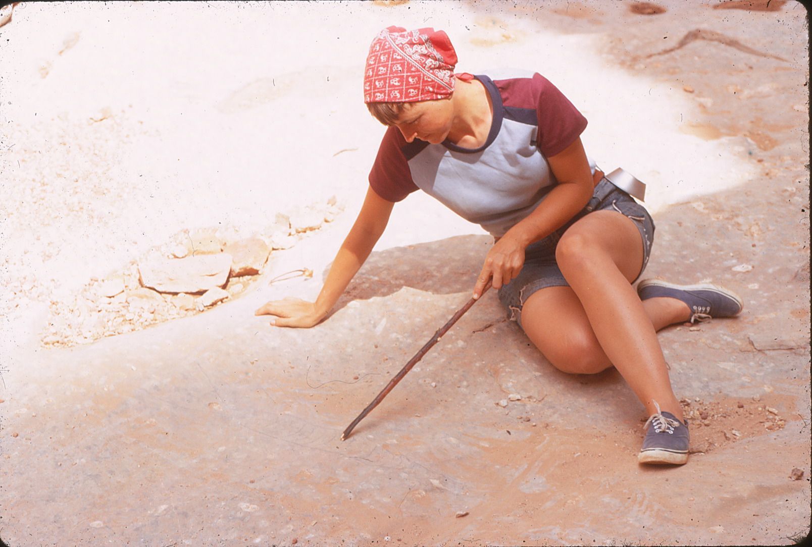 Woman kneeling, examining ground with a stick. Wearing a red bandanna, blue and red shirt, denim shorts, and sneakers.