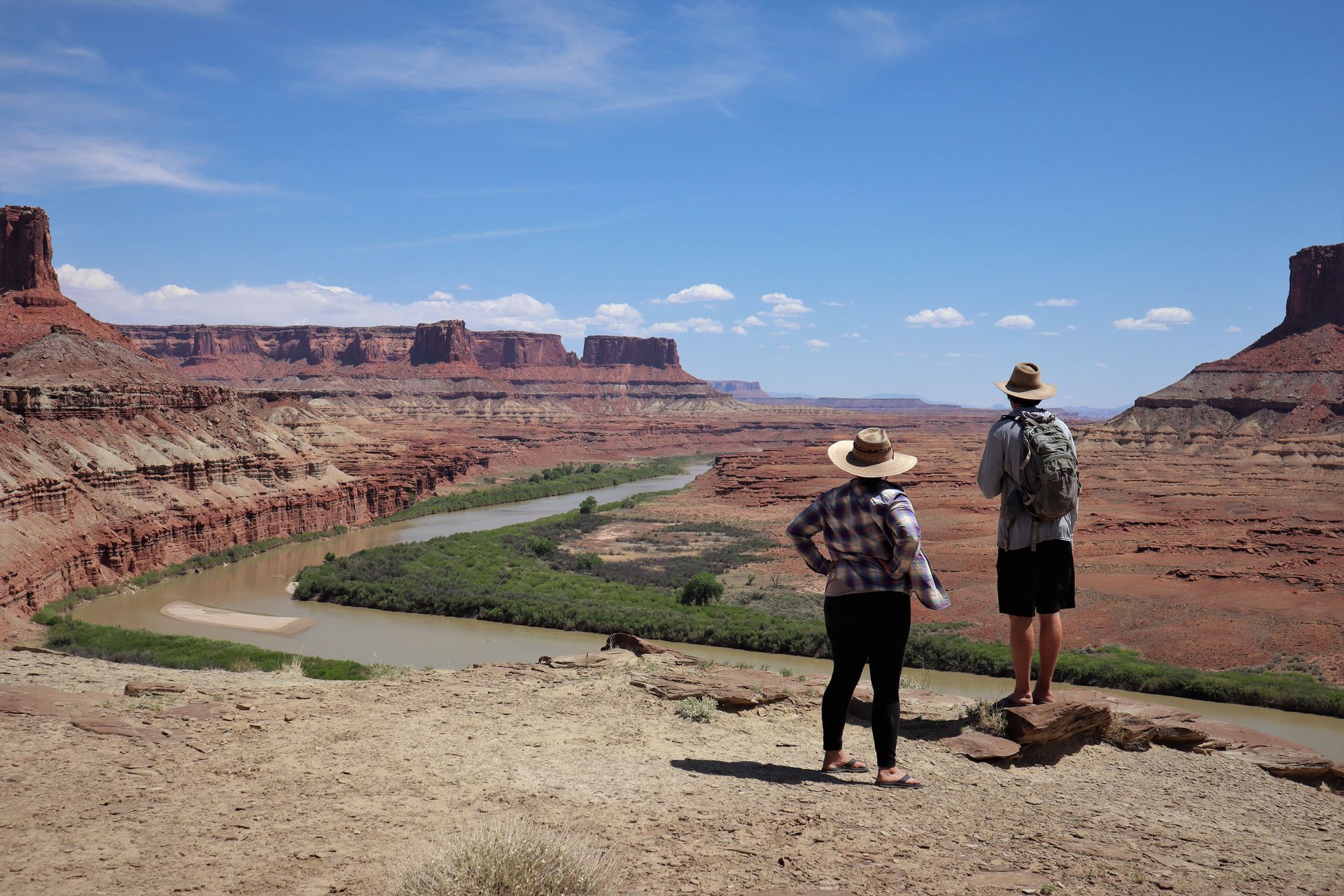 Two people look over a canyon, river below. Red rock formations under blue sky.