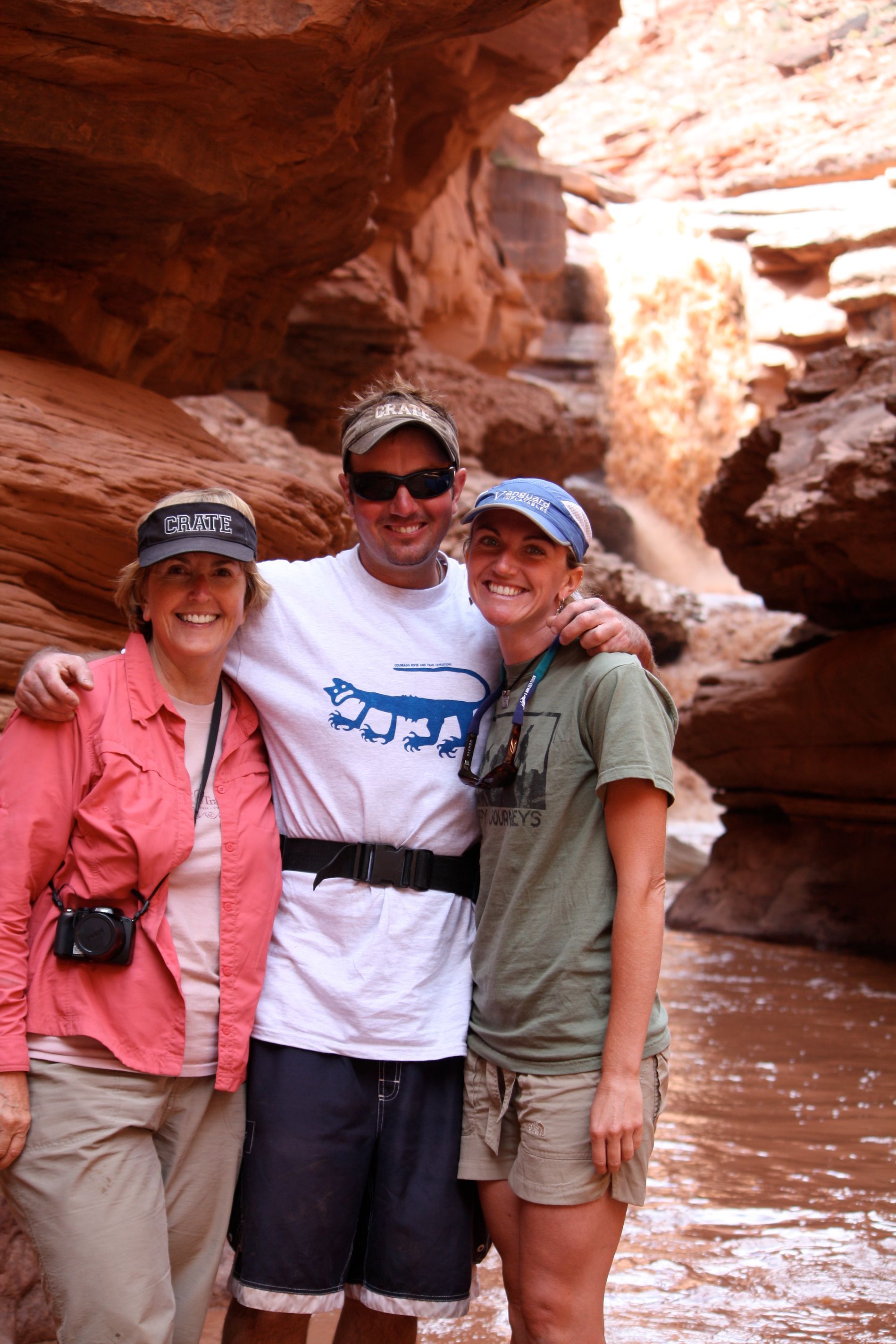 Three people pose in a narrow canyon with red rock walls. They smile, arms around each other.