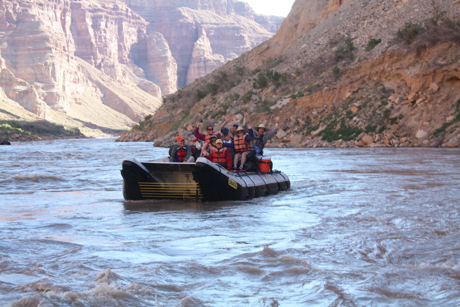 A raft capsizes in a turbulent river, spraying water against a red-brown rock face.
