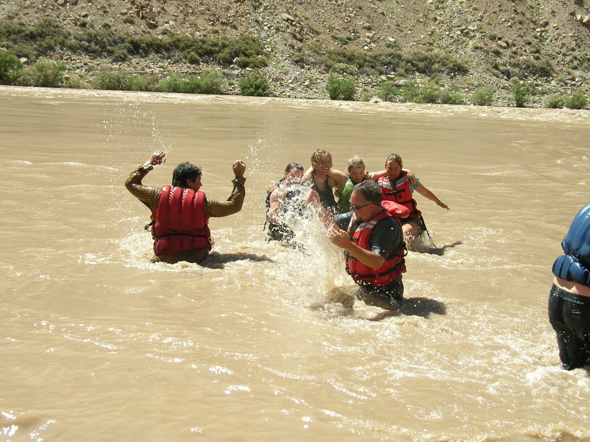 People splashing in brown river water, wearing life vests, near a rocky bank.