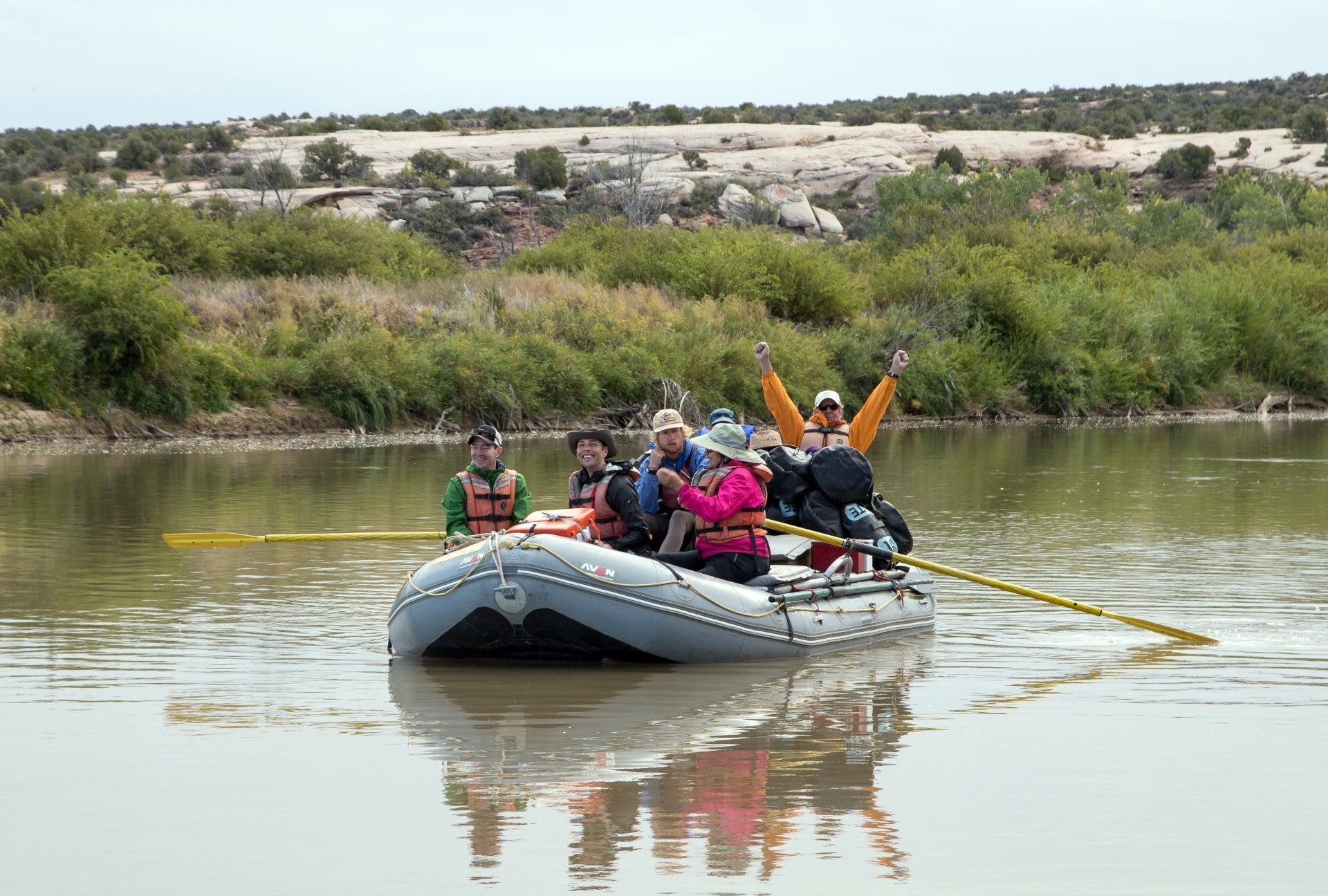 People on a raft with paddles, on a river with green bushes in background. Cloudy sky.