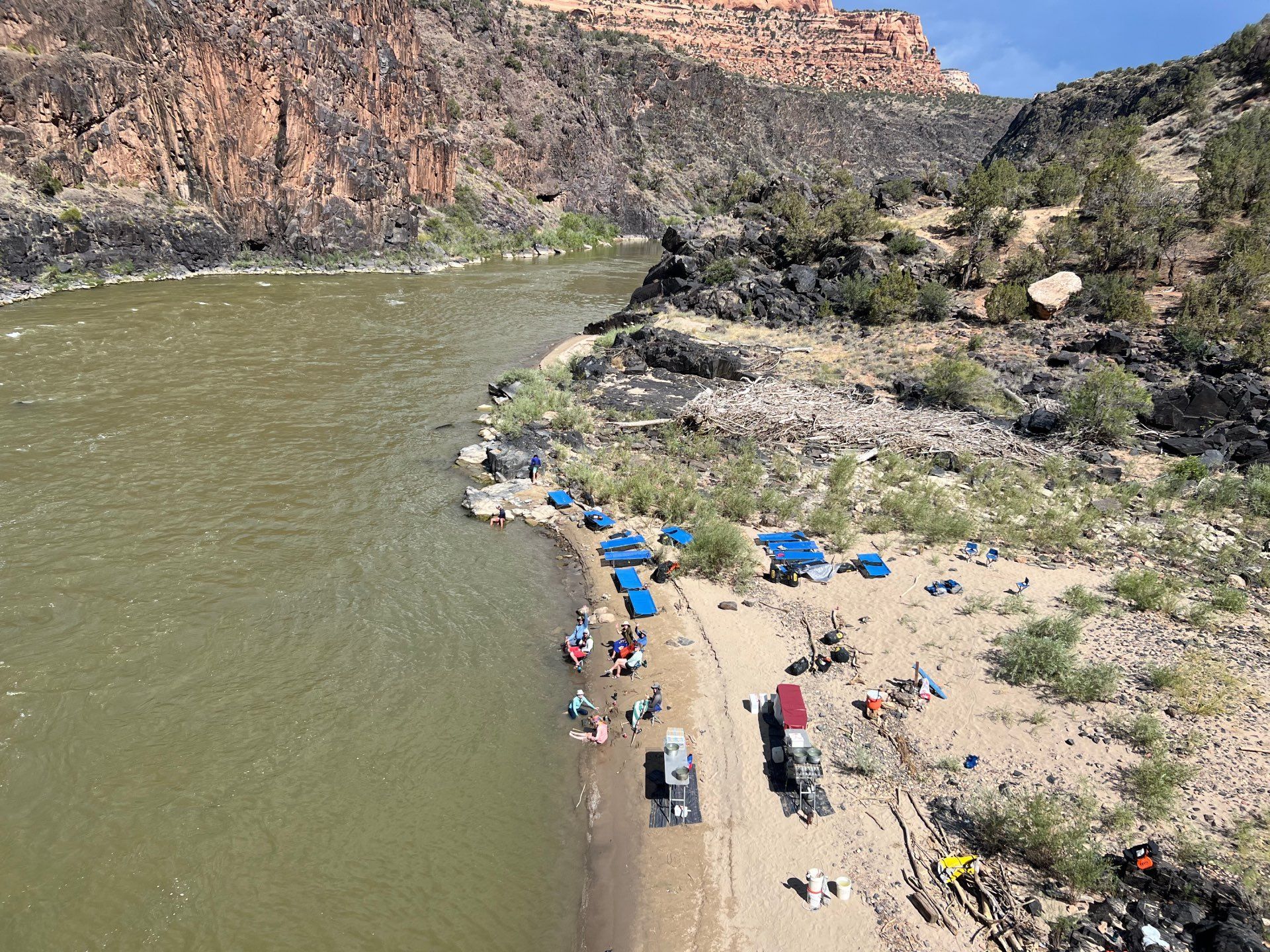 Riverbank with tents, people, and vehicles, alongside a brown river and cliffs.