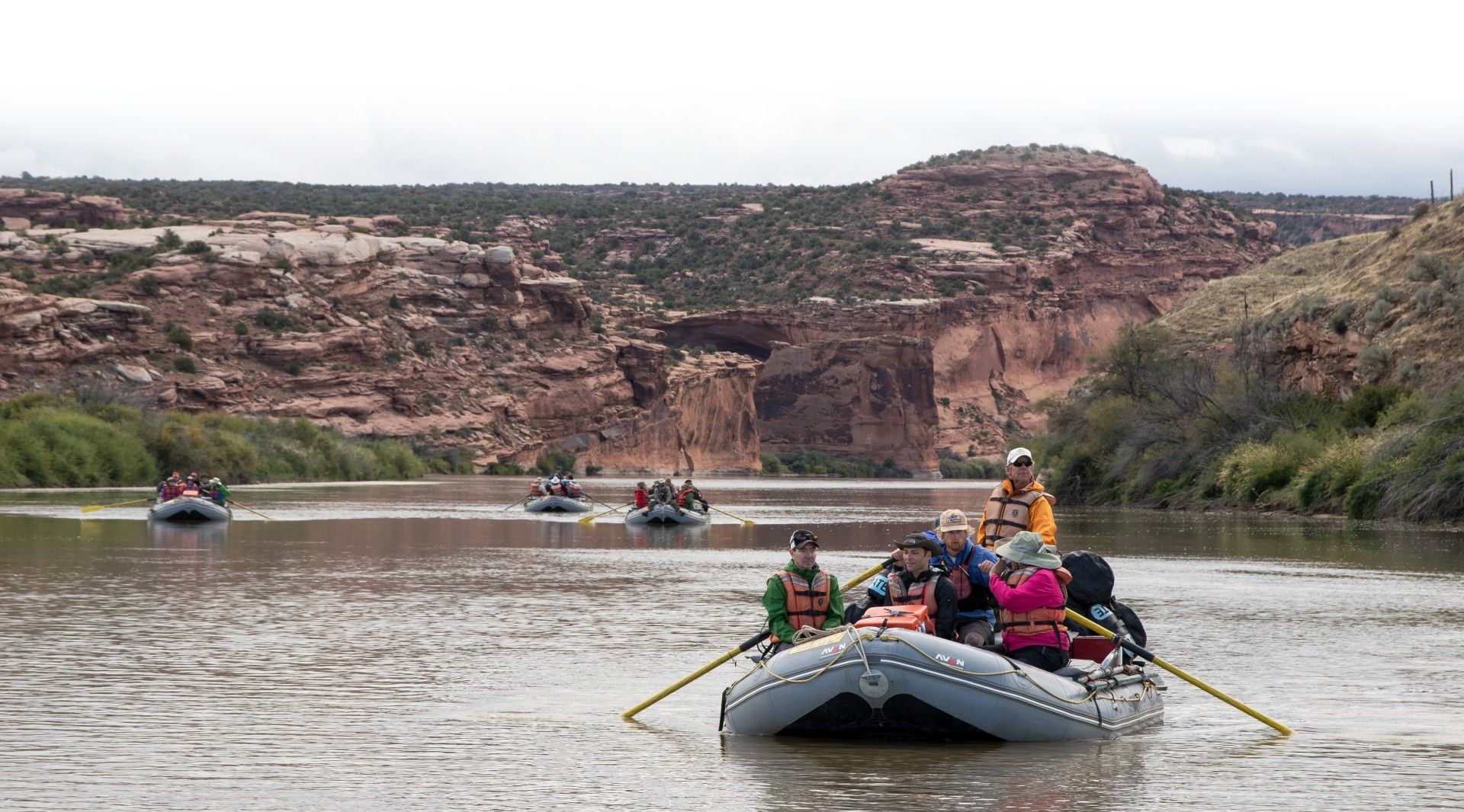Several rafts with people on a river, cliffs in the background, cloudy sky.